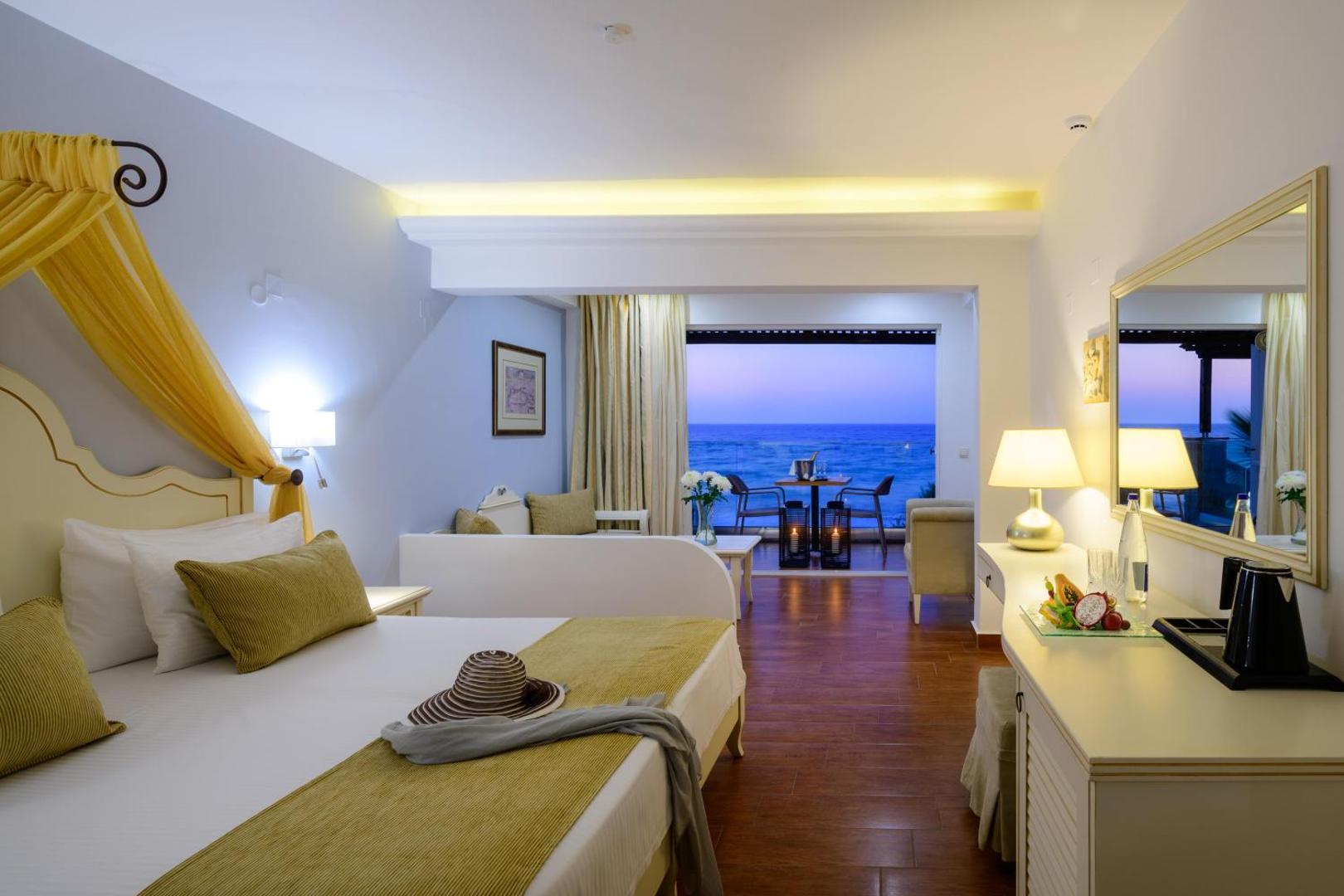 Alexander Beach Hotel & Village mājīgs numurs ar skatu uz jūru no balkona, gaišām mēbelēm un dzelteniem akcentiem interjerā.