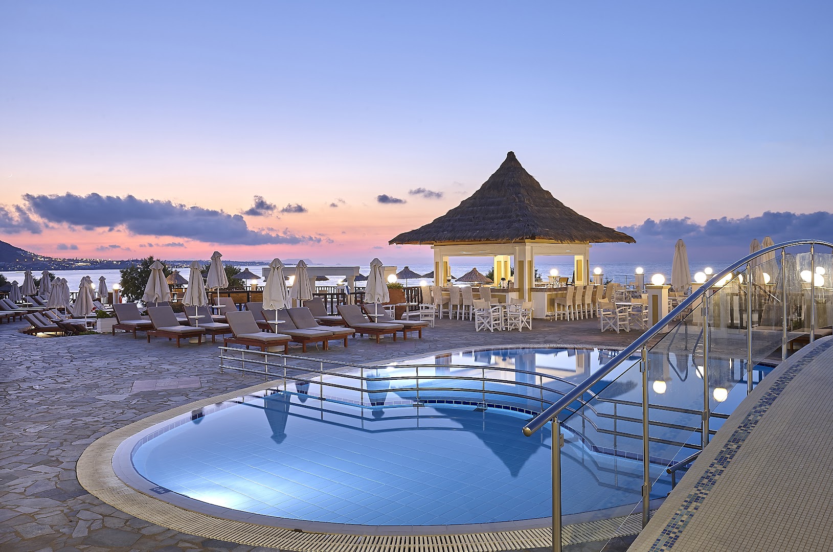Alexander Beach Hotel & Village āra baseins un bārs saulrieta laikā Krētā ar skatu uz jūru un kalniem tālumā