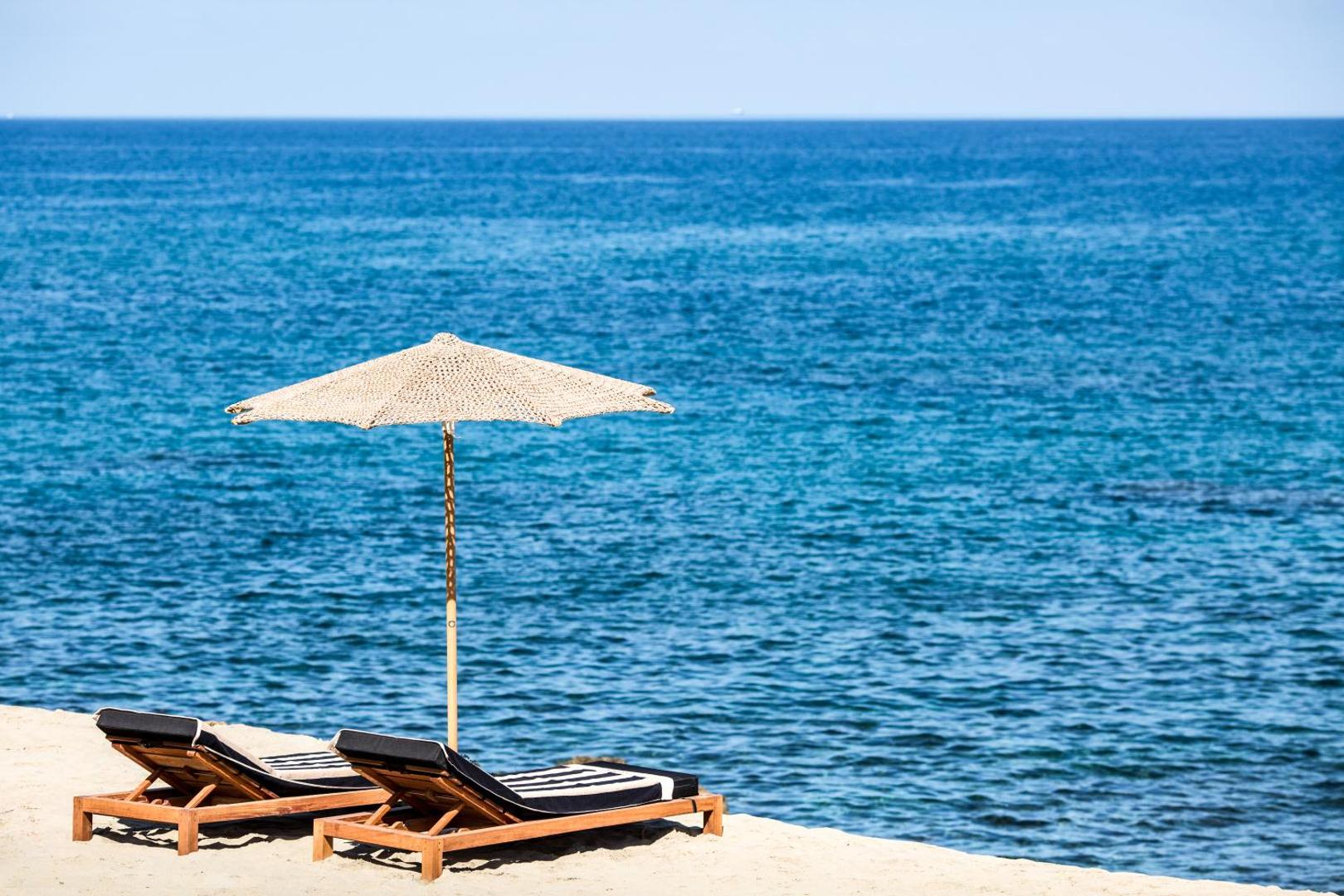 Abaton Island Resort & Spa privātā pludmale Krētā ar diviem koka sauļošanās krēsliem un saulessargu uz gaišām smiltīm pie zilas jūras