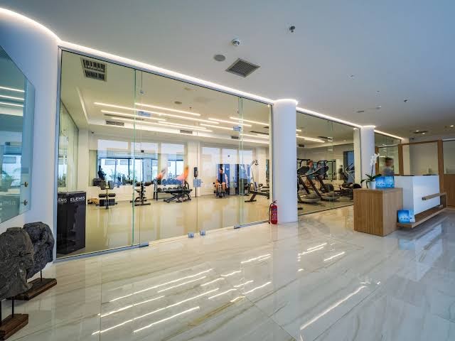 Abaton Island Resort & Spa moderns fitness centrs ar stikla sienām, trenažieriem un gaišu marmora grīdu viesnīcas spa zonā
