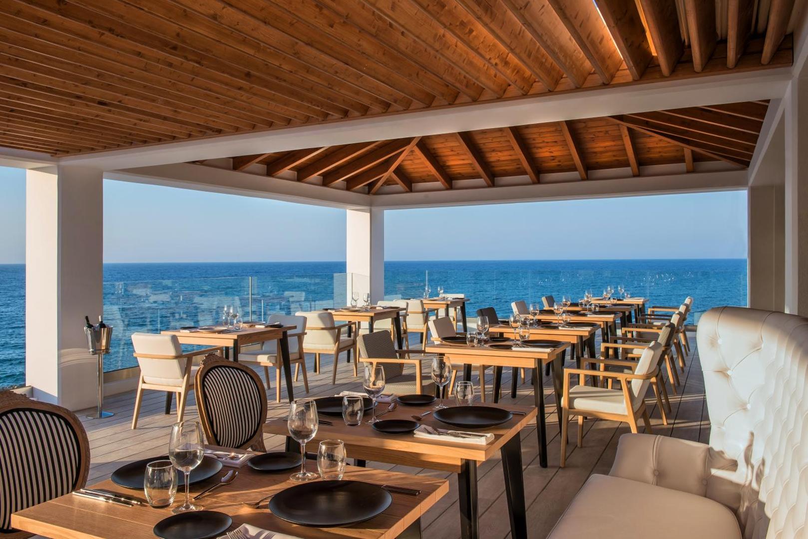 Abaton Island Resort & Spa restorāna terase ar koka griestiem un skatu uz jūru, eleganti klāti galdi vakariņām piekrastē