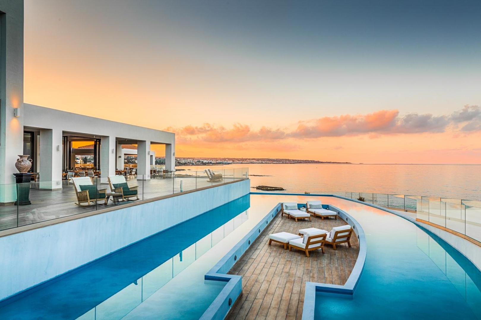 Abaton Island Resort & Spa āra terase un baseins saulrietā Krētā — skats uz jūru un piekrastes pilsētu tālumā