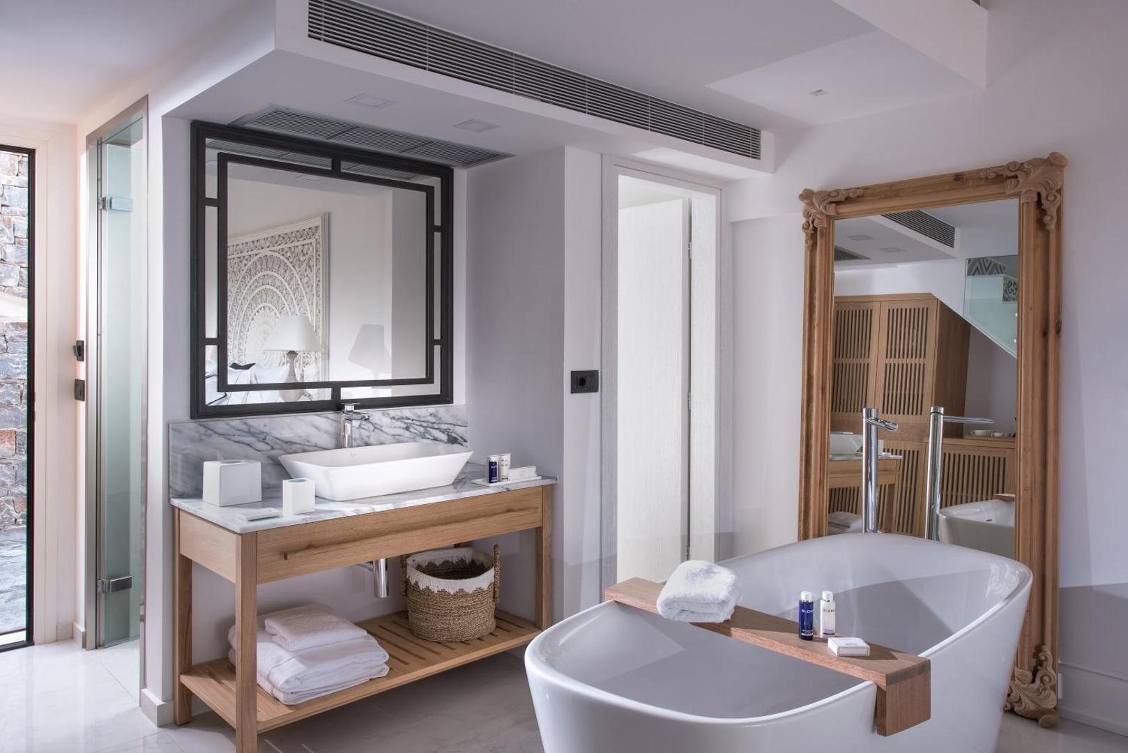 Abaton Island Resort & Spa luksusa numura vannasistaba ar brīvstāvošu vannu, marmora virsmām un lielu spoguli kokgriezuma rāmī.