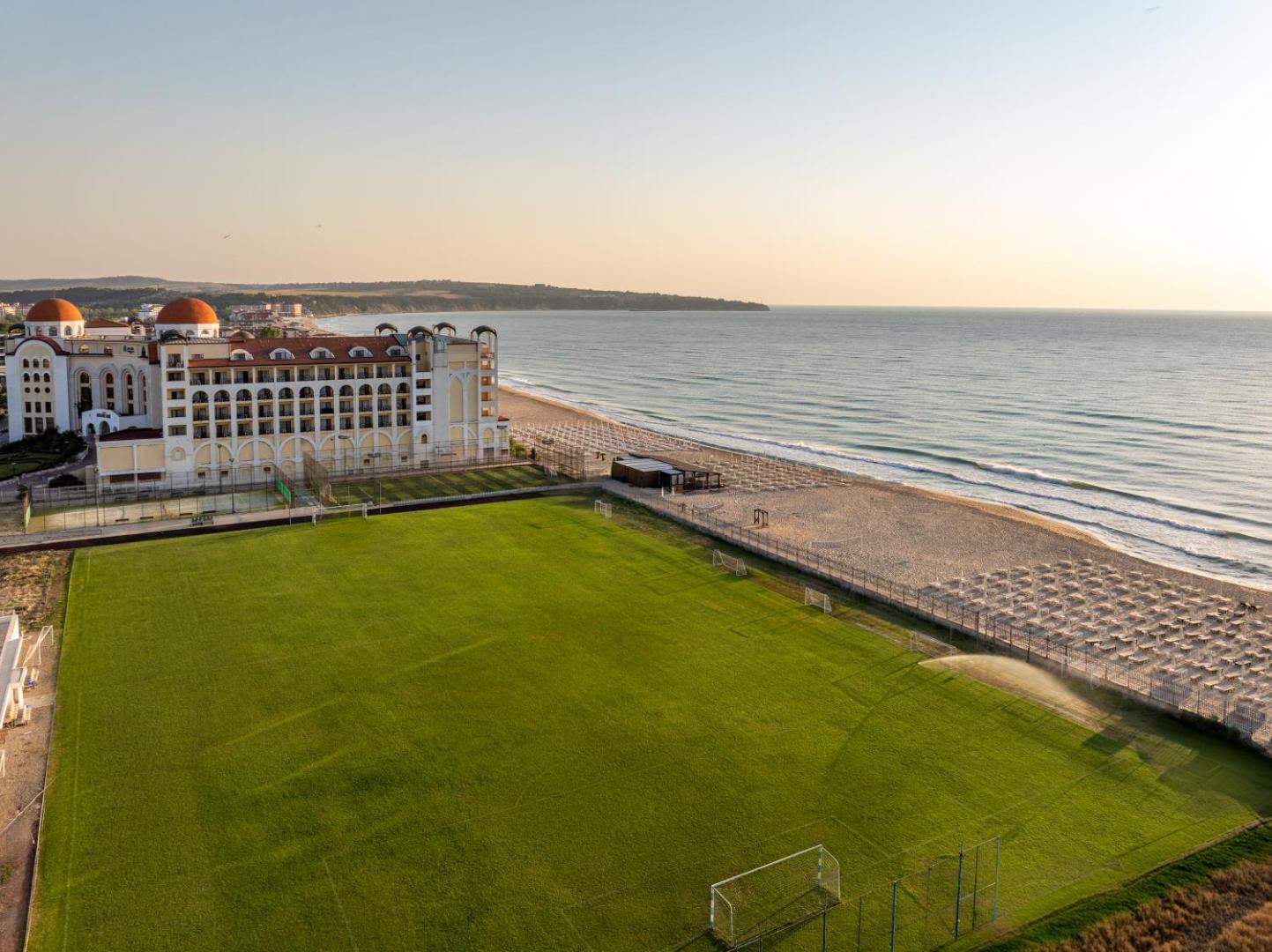 Alua Helios Bay futbola laukums ar dabisko zālāju blakus plašajai smilšu pludmalei un viesnīcas galvenajai ēkai saulrietā