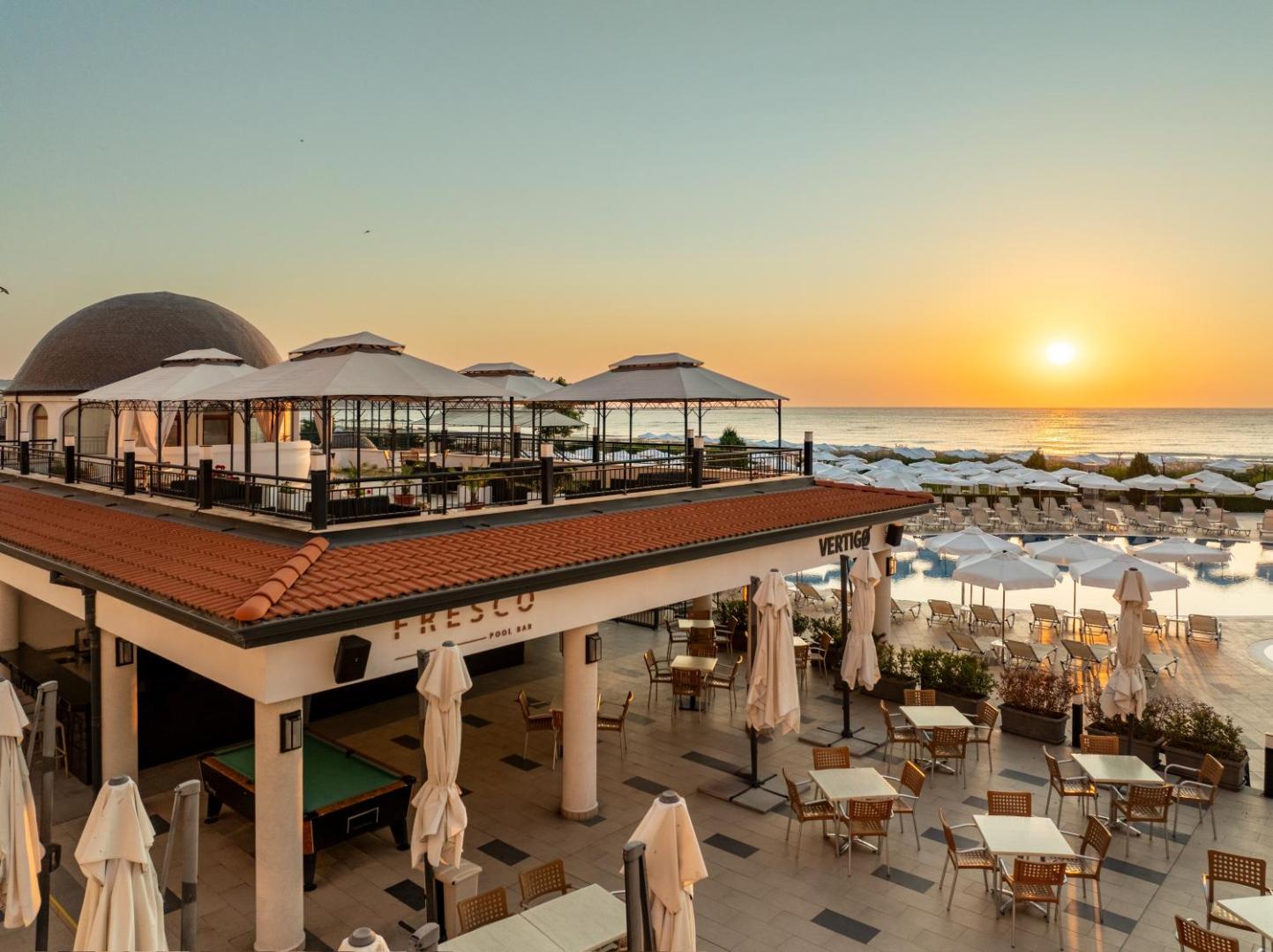 Alua Helios Bay āra terase un baseina zona saulrietā — skats uz Fresco bāru, saulessargiem un jūras piekrasti Obzorā