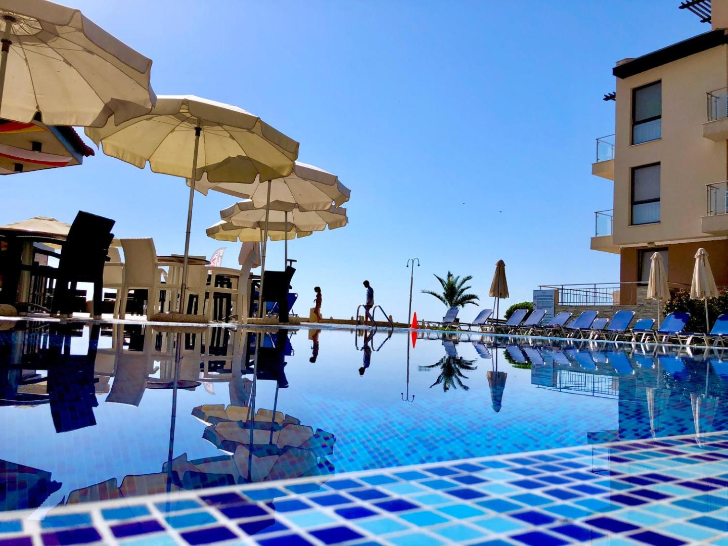 Obzor Beach Resort baseina ūdens atspulgs ar saulessargiem un viesnīcas ēku saulainā dienā Obzorā, Bulgārijā
