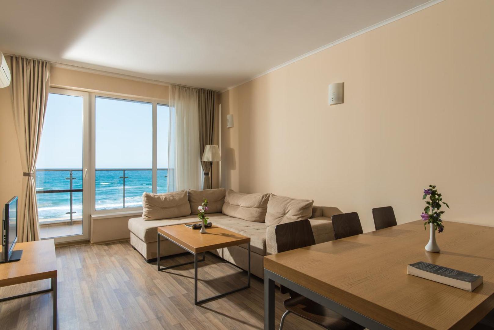 Mājīgs Obzor Beach Resort numurs ar stūra dīvānu, ēdamgaldu un tiešu skatu uz jūras viļņiem caur logu