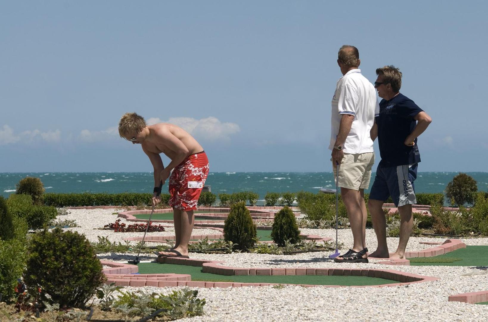 Viesi spēlē minigolfu Obzor Beach Resort teritorijā pie jūras, saulains laiks un zaļš mākslīgā seguma laukums
