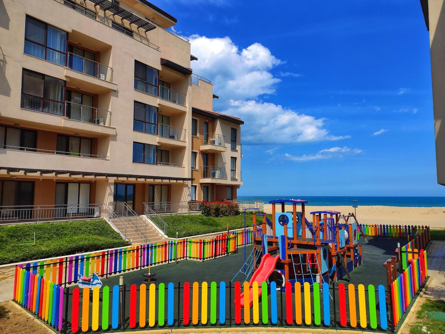 Obzor Beach Resort bērnu rotaļu laukums ar krāsainu sētu pie viesnīcas fasādes un pludmales fona