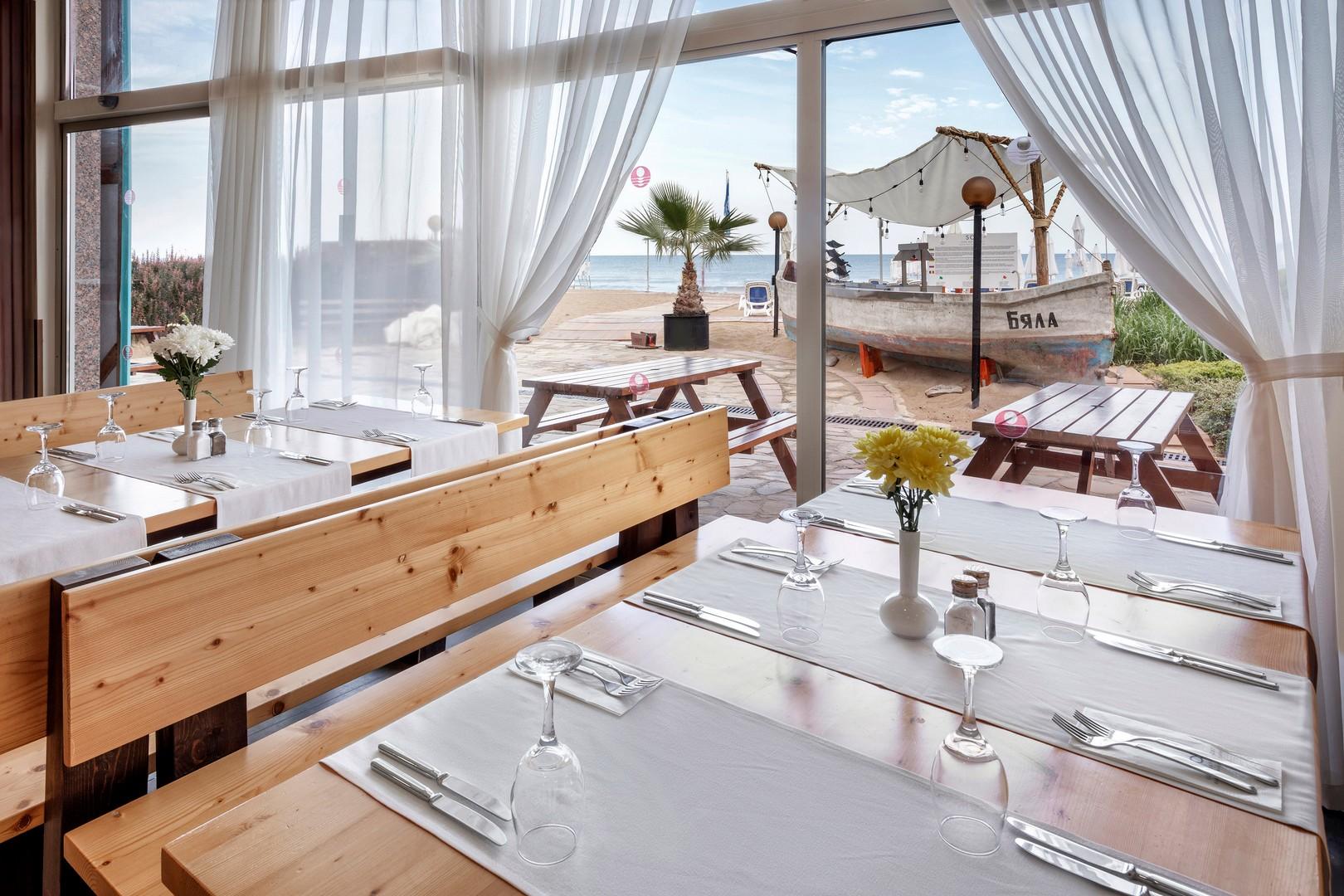 Sol Luna Bay Resort restorāna terase ar koka galdiem un skatu uz pludmali, kurā redzama dekoratīva zvejnieku laiva.