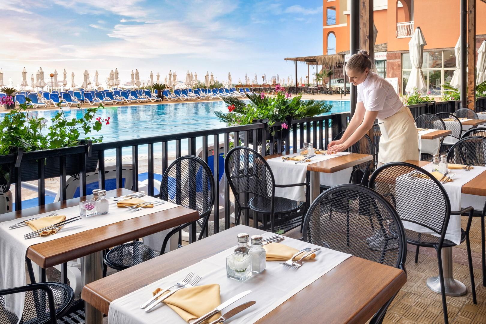 Sol Luna Bay Resort āra terase pie baseina ar klātiem galdiem un viesmīli, fonā redzama pludmales zona ar saulessargiem.