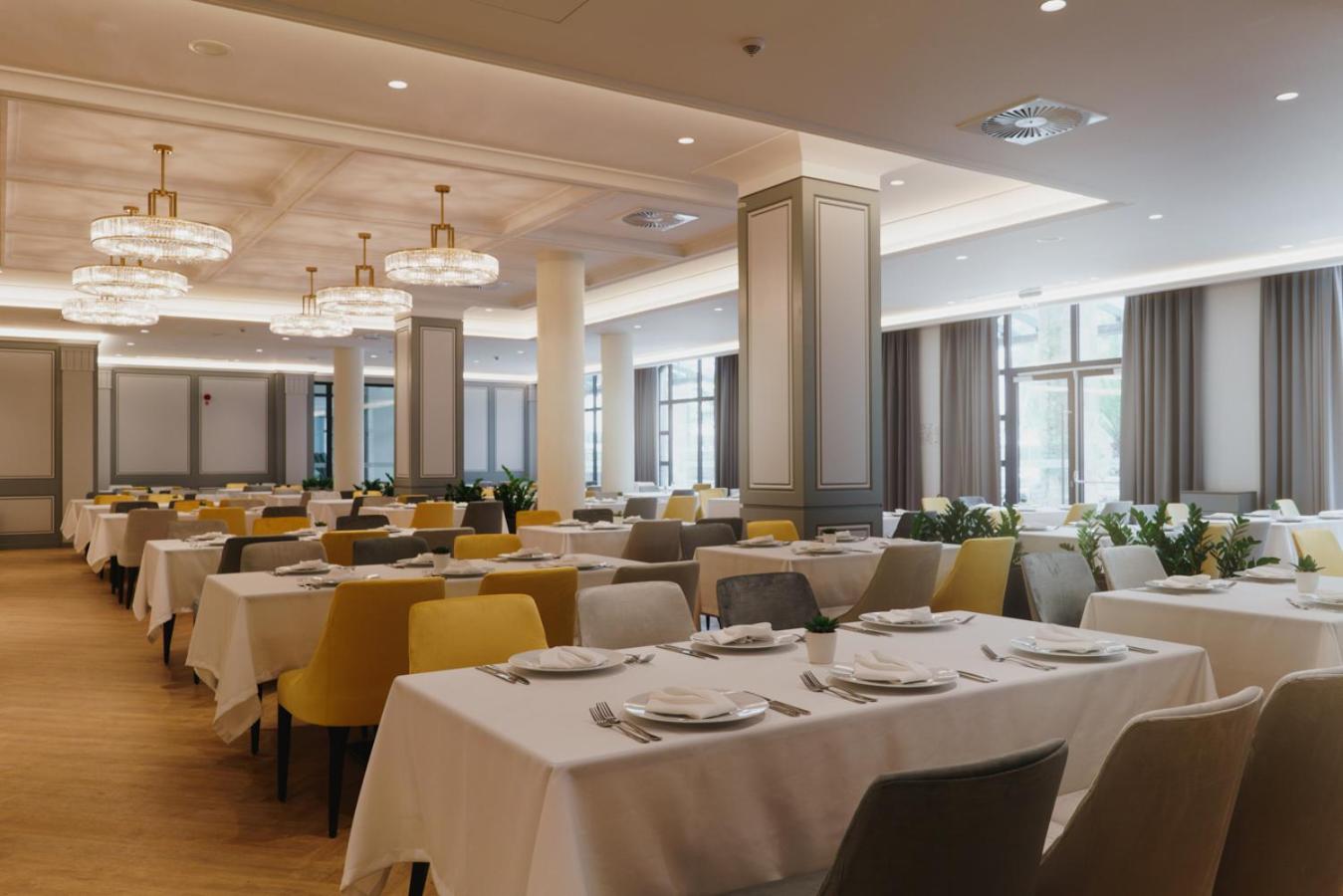 Astor Garden viesnīcas plašais restorāns ar elegantiem galdiem, dzelteniem krēsliem un modernām lustrām gaišā interjerā