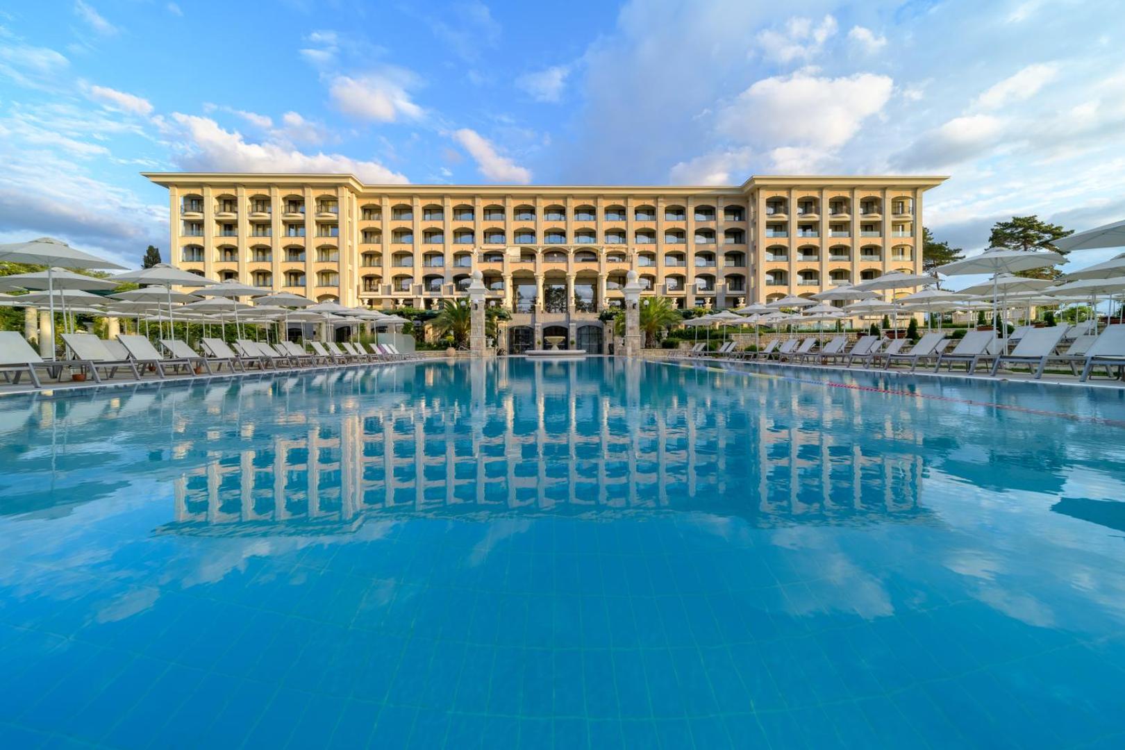 Panorāmas skats uz Astor Garden viesnīcas ēku un zilo āra baseinu Bulgārijā, ko ieskauj balti saulessargi un atpūtas zona.