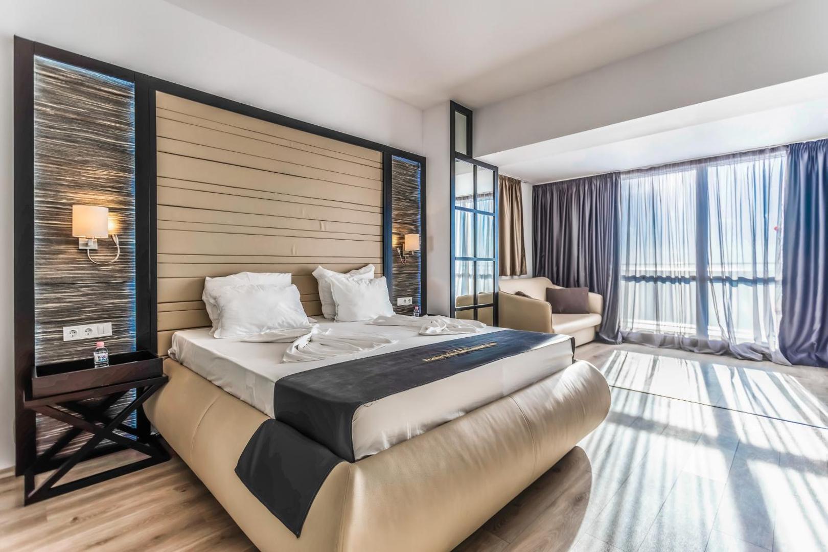 Prestige Deluxe Hotel Aquapark Club gaišs viesnīcas numurs ar lielu gultu, dīvānu un panorāmas logiem, kas nodrošina dabisku apgaismojumu