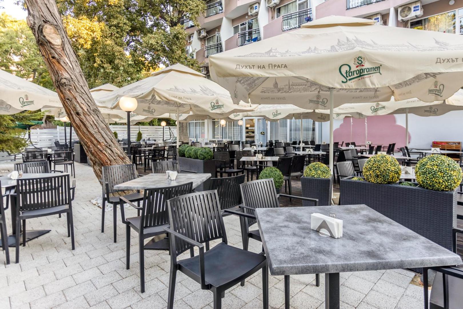 Prestige Deluxe Hotel Aquapark Club āra terase ar galdiņiem un saulessargiem Zelta Smiltīs, Bulgārijā — mājīga atpūtas zona koku ēnā
