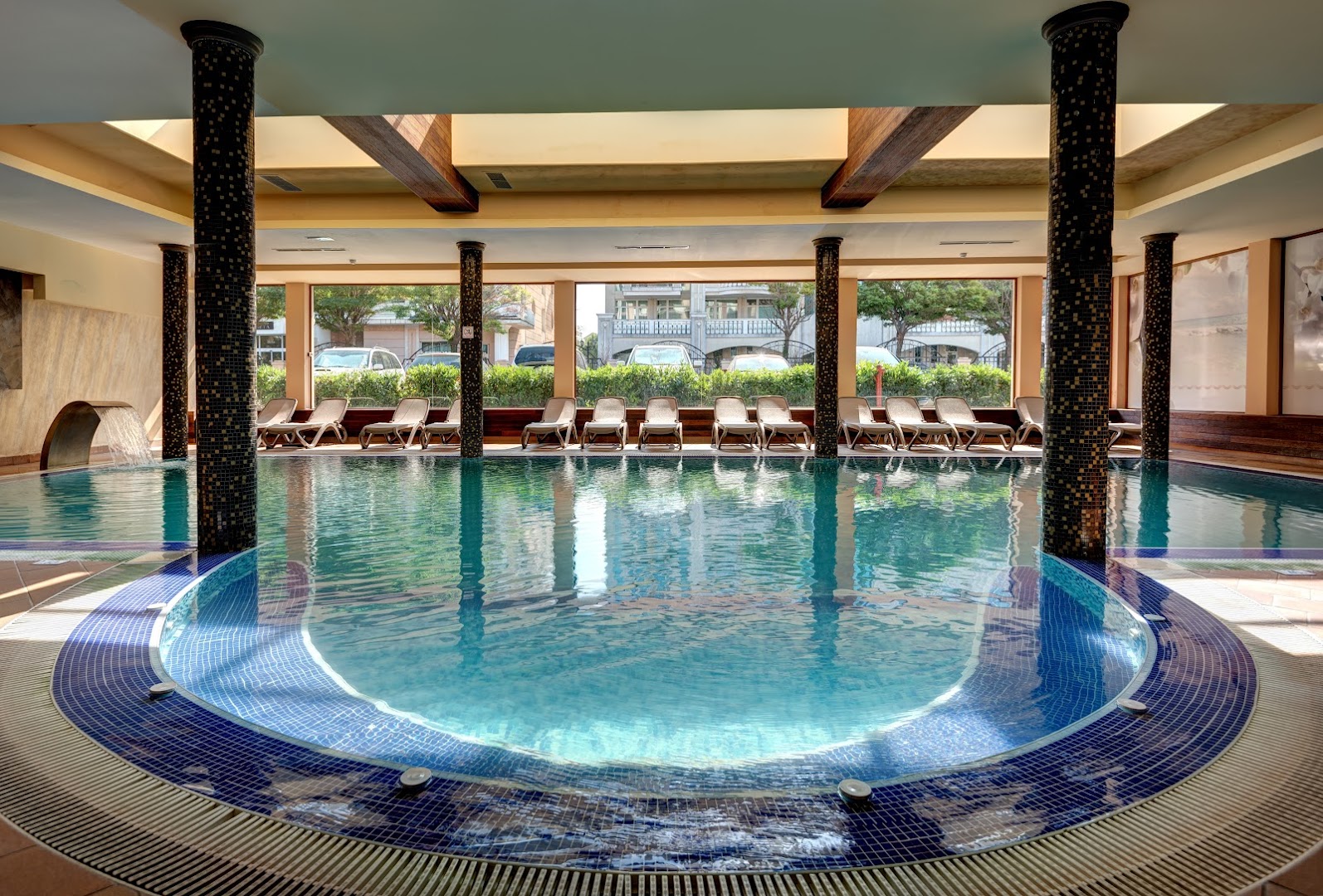 Festa Via Pontica Resort iekštelpu baseins ar kolonnām un atpūtas krēsliem spa centrā Pomorijā — plaša relaksācijas zona