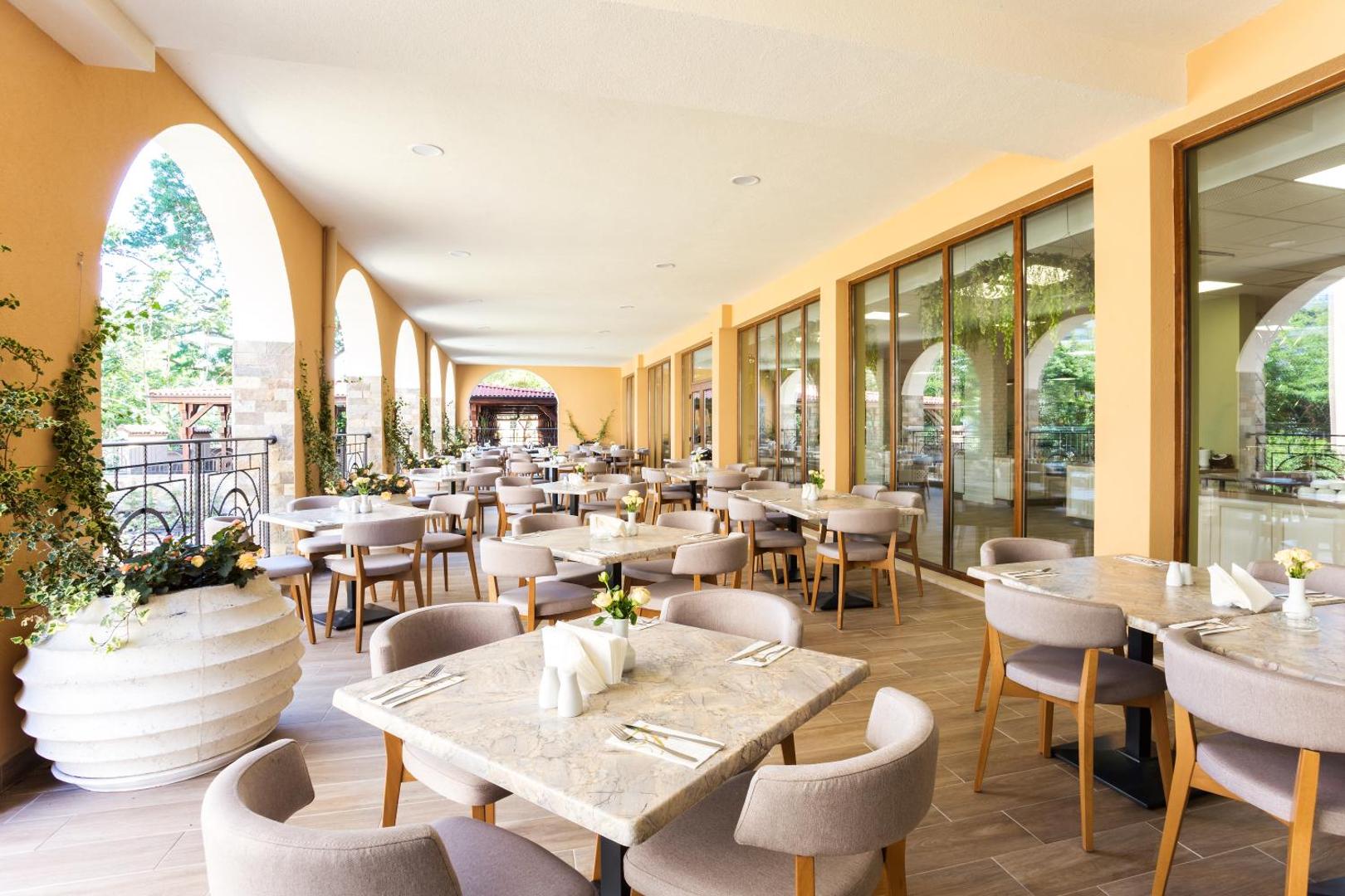 Festa Via Pontica Resort plašā restorāna terase ar marmora galdiem un skatu uz viesnīcas dārzu un arkveida arhitektūru.