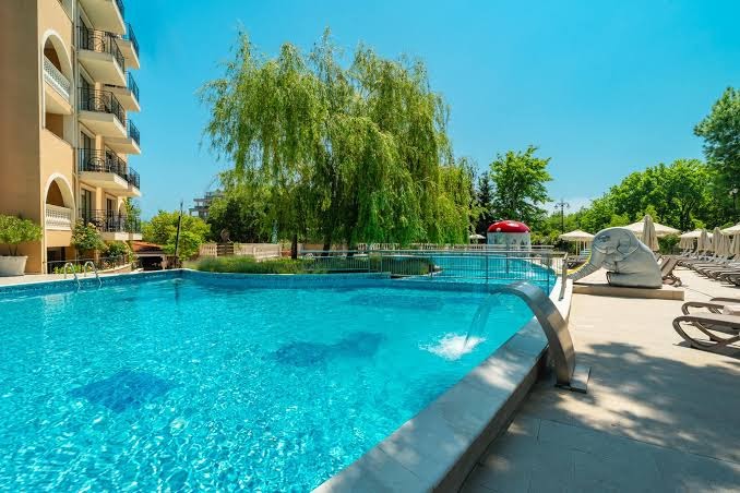 Festa Via Pontica Resort āra baseins ar kaskādi un bērnu rotaļu elementiem saulainā dienā Pomorijā — apkārt zaļojoši koki