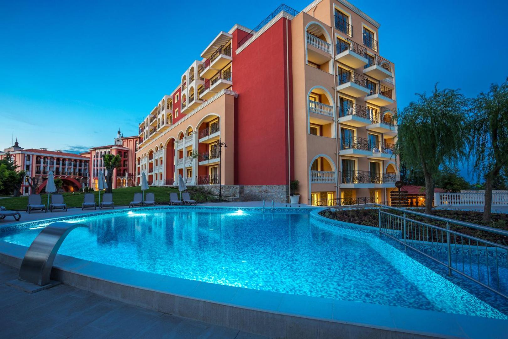 Festa Via Pontica Resort āra baseins vakarā ar izgaismotu viesnīcas fasādi un sauļošanās krēsliem zālienā