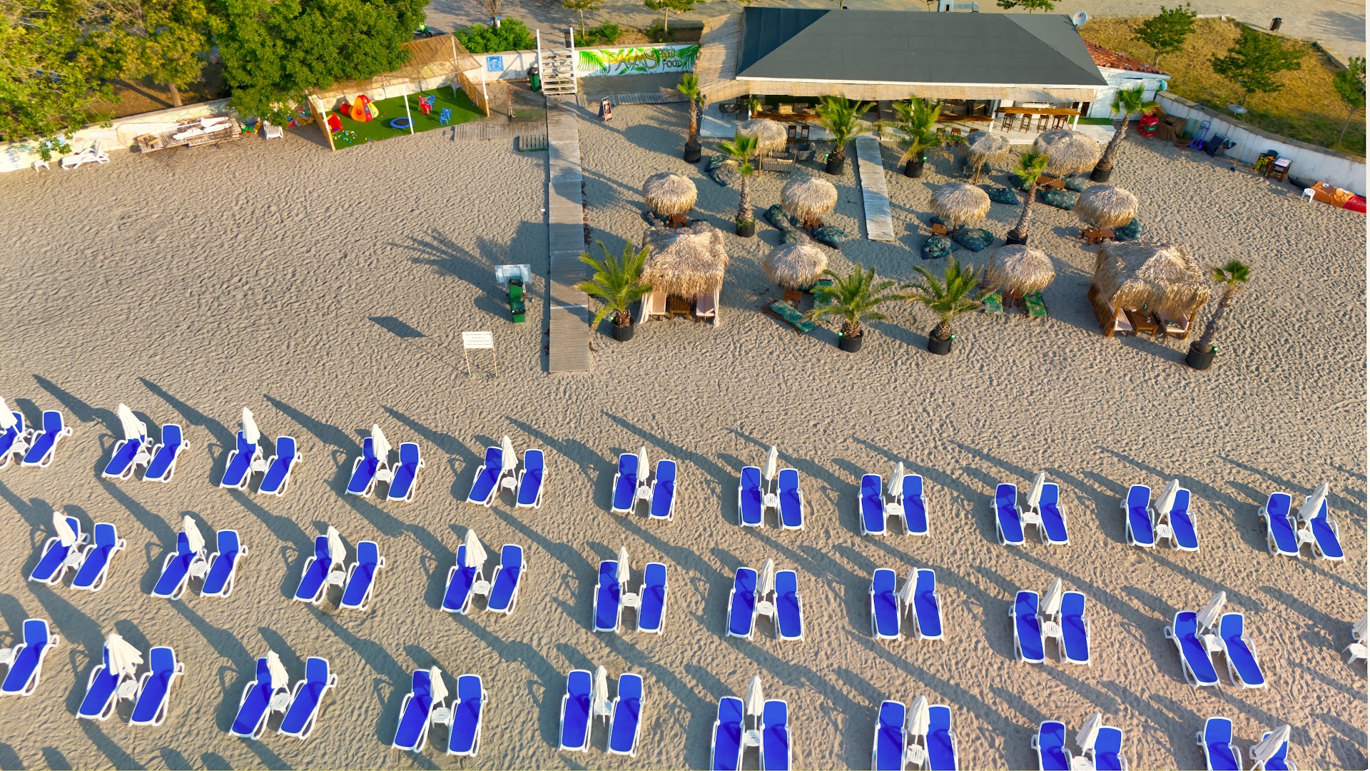 Festa Via Pontica Resort privātā pludmale Pomorijā no putna lidojuma ar ziliem sauļošanās krēsliem, saulessargiem un pludmales bāru.