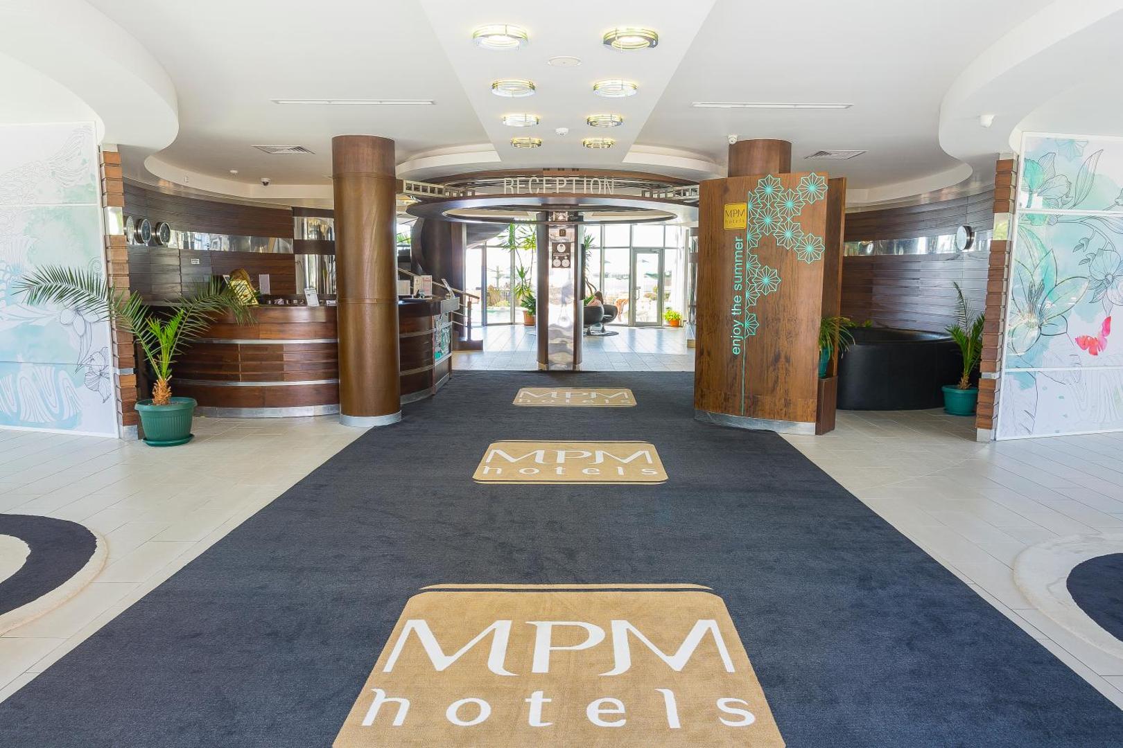 MPM Hotel Arsena plašā un gaišā recepcijas zona ar zīmola paklājiem, koka elementiem un ieeju viesnīcas vestibilā