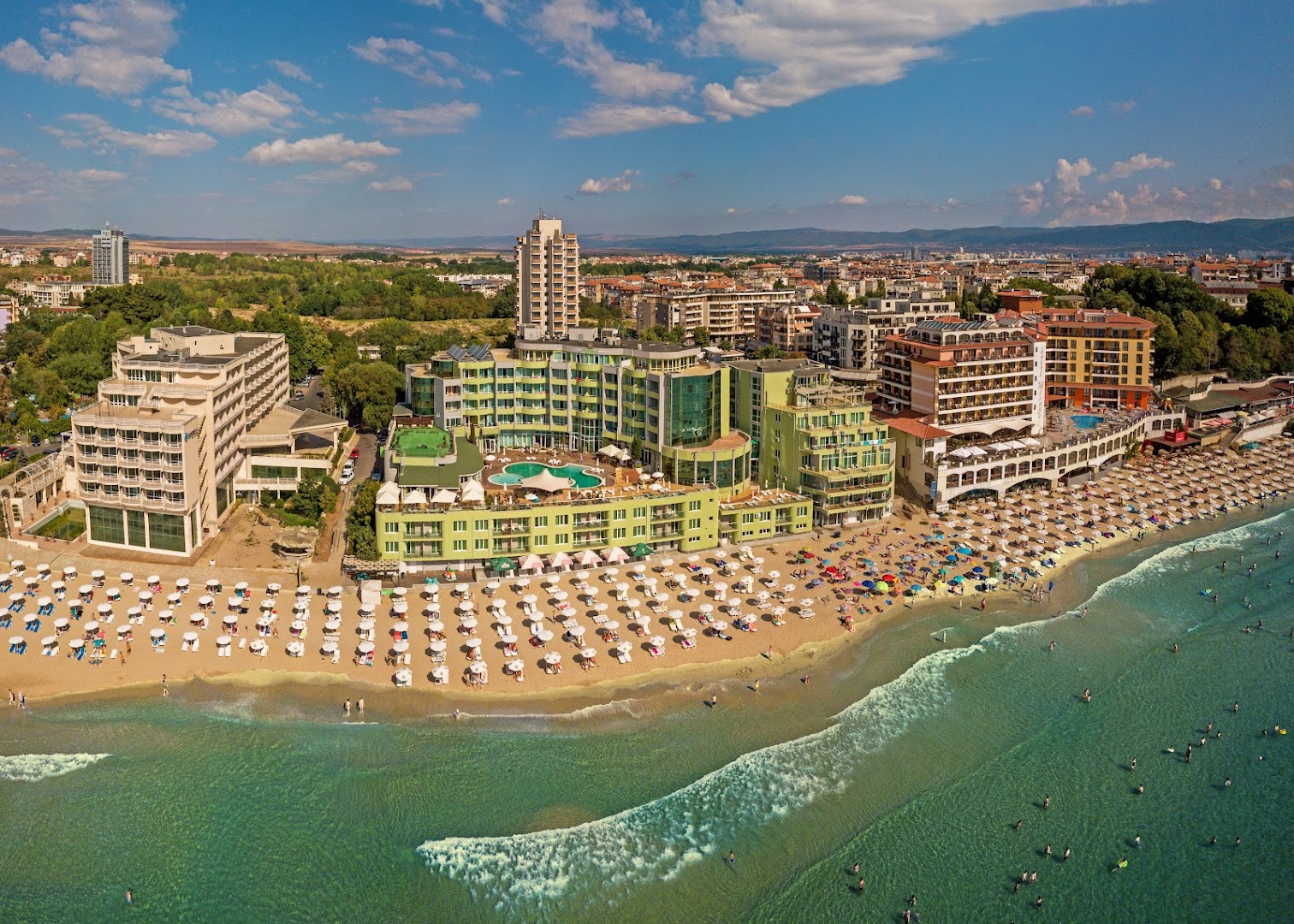 MPM Hotel Arsena skats no putna lidojuma Nesebārā, Bulgārijā — viesnīcas ēka tieši pie smilšu pludmales ar saulessargiem un jūras krastu
