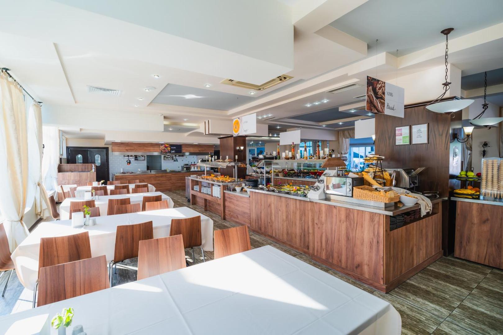 MPM Hotel Arsena galvenais restorāns ar bufetes līniju, koka apdari un gaišiem galdiem — plaša brokastu un vakariņu zona