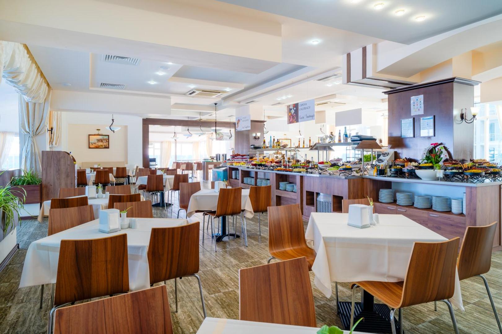 MPM Hotel Arsena restorāna iekštelpas ar bufetes tipa ēdināšanas zonu un saklātiem galdiem gaišā interjerā