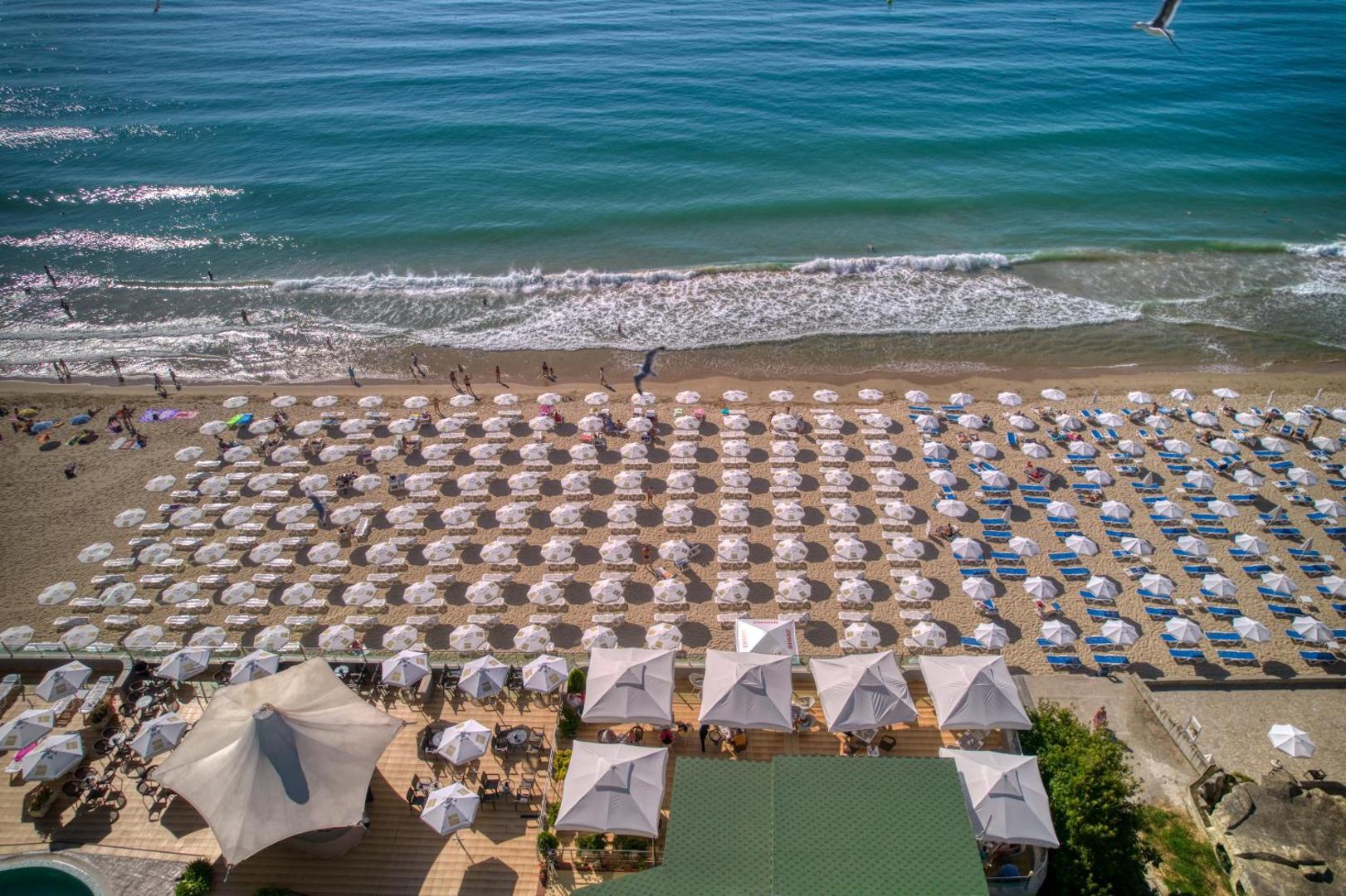 MPM Hotel Arsena privātā pludmale Nesebārā no putna lidojuma — simtiem saulessargu un sauļošanās krēslu pie dzidri zilās Melnās jūras