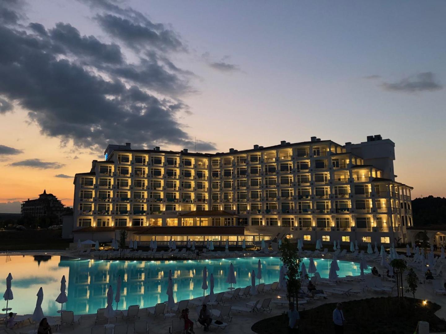 Sunrise Blue Magic Resort viesnīcas ēka saulrieta laikā ar izgaismotiem numuriem un baseina atspulgu vakarā