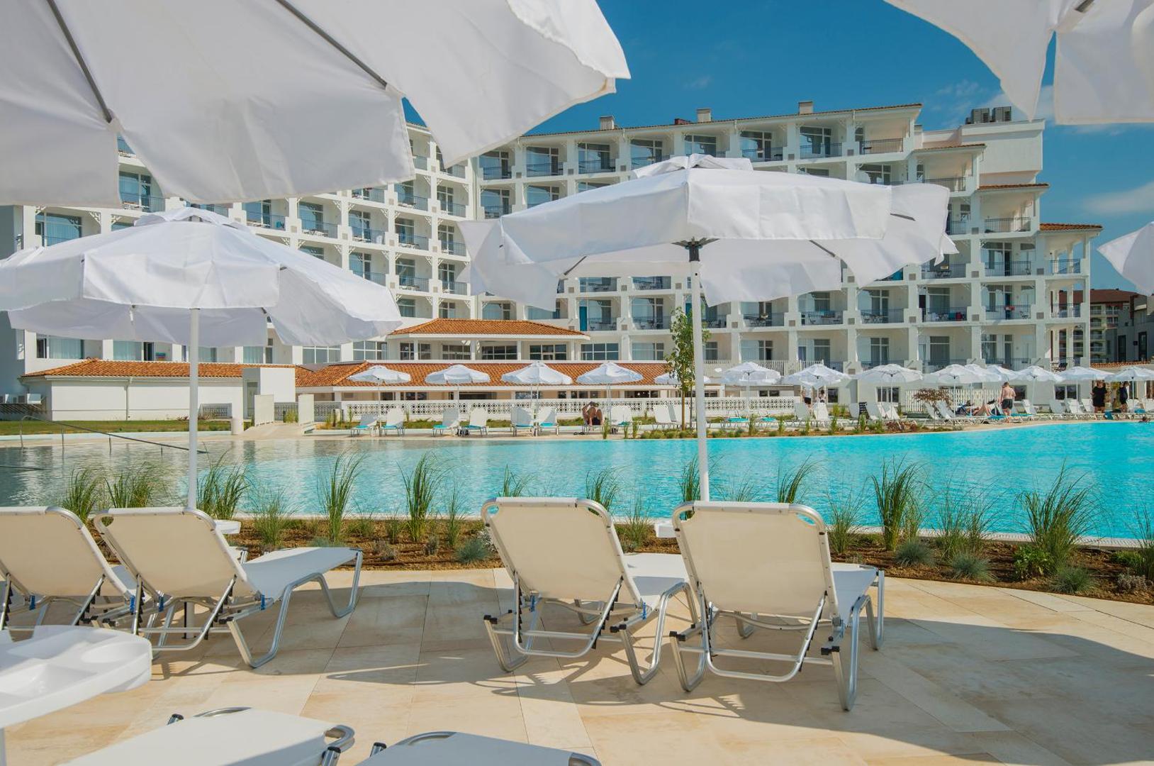 Atpūtas krēsli un balti saulessargi pie Sunrise Blue Magic Resort baseina ar skatu uz viesnīcas galveno ēku saulainā dienā