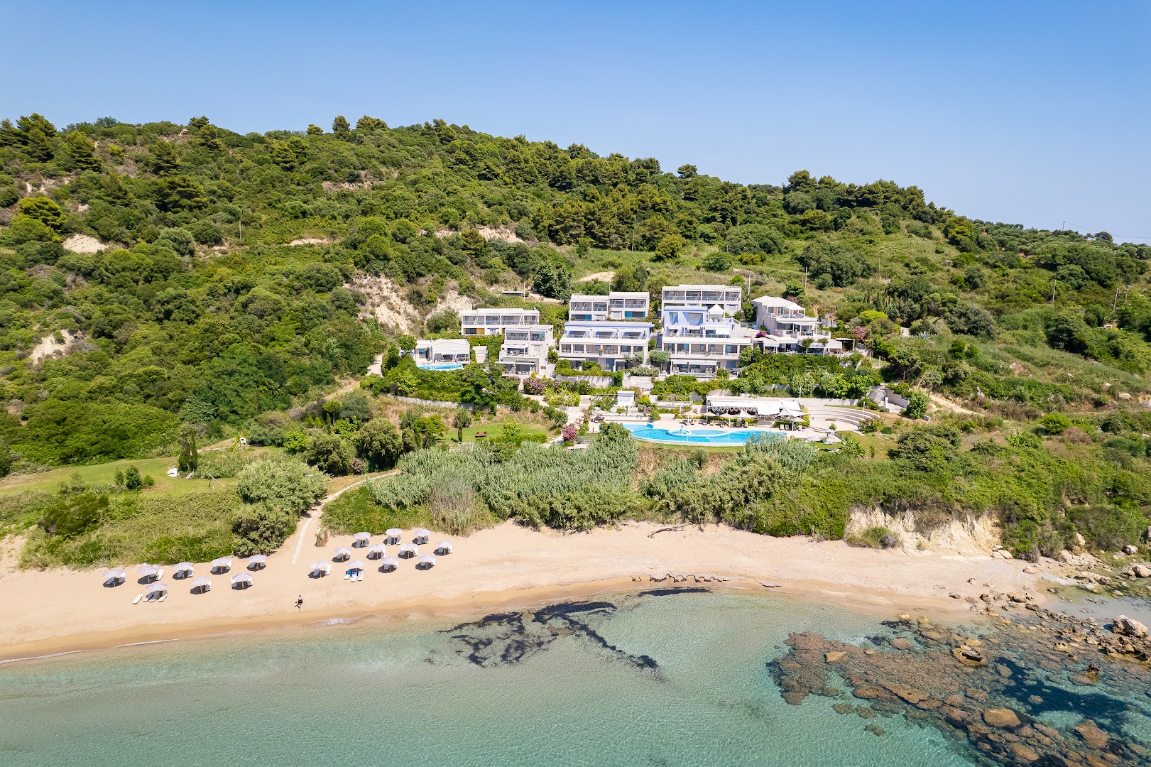 Mare Dei Ionian Resort skats no jūras puses uz viesnīcas ēkām kalna nogāzē, baseinu un smilšu pludmali ar saulessargiem