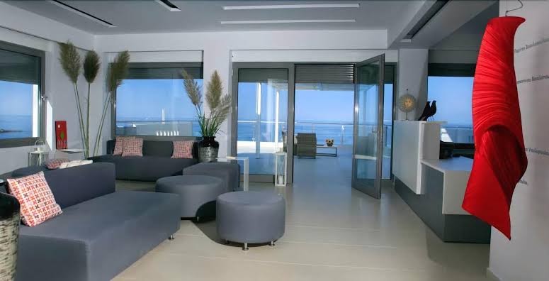 Mare Dei Ionian Resort dizaina lobby telpa ar pelēkiem dīvāniem, dekoratīviem elementiem un izeju uz terasi ar skatu uz jūru.
