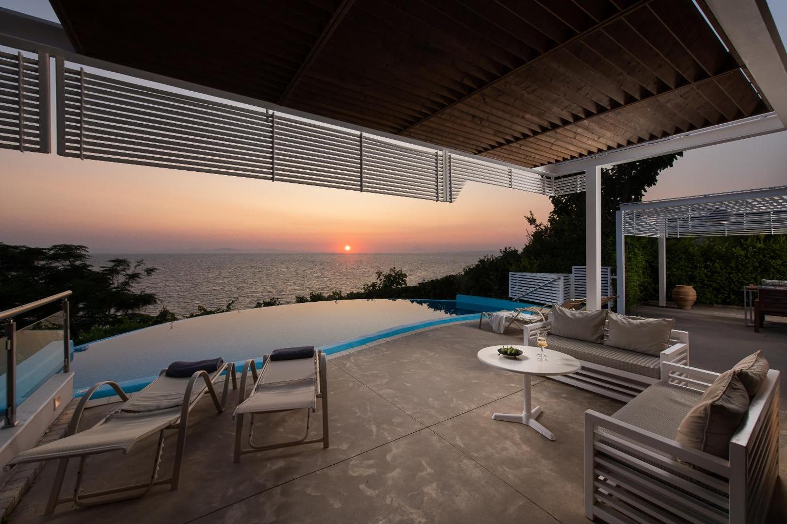 Mare Dei Ionian Resort terase ar skatu uz saulrietu Jonijas jūrā — atpūtas zona ar dīvāniem un baseinu piekrastē