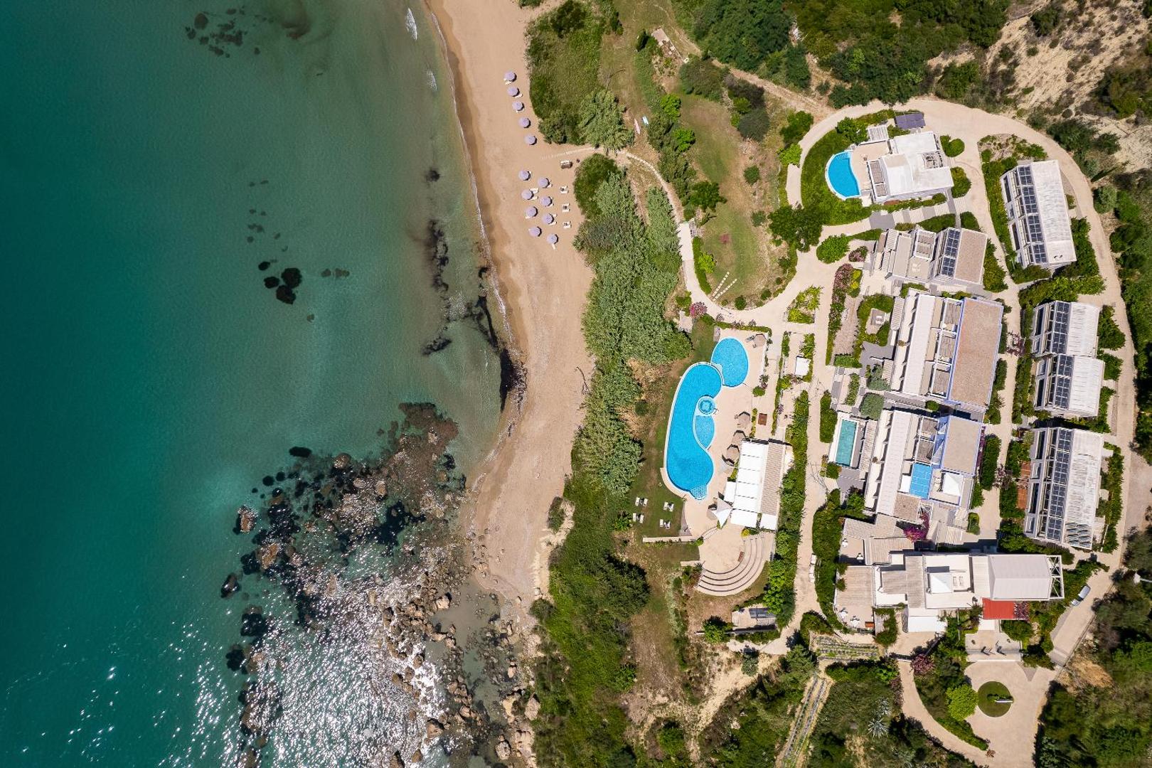 Mare Dei Ionian Resort aerofoto skats tieši no augšas uz baseinu zonu, pludmali ar saulessargiem un viesnīcas ēku izvietojumu dārzā