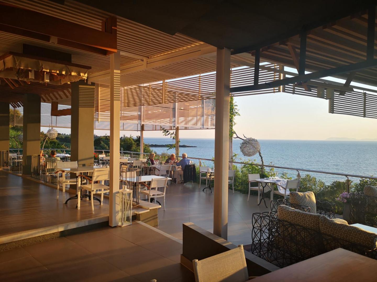 Mare Dei Ionian Resort restorāna āra terase ar galdiņiem un skatu uz jūru saulrieta laikā Peloponēsas reģionā, Grieķijā.