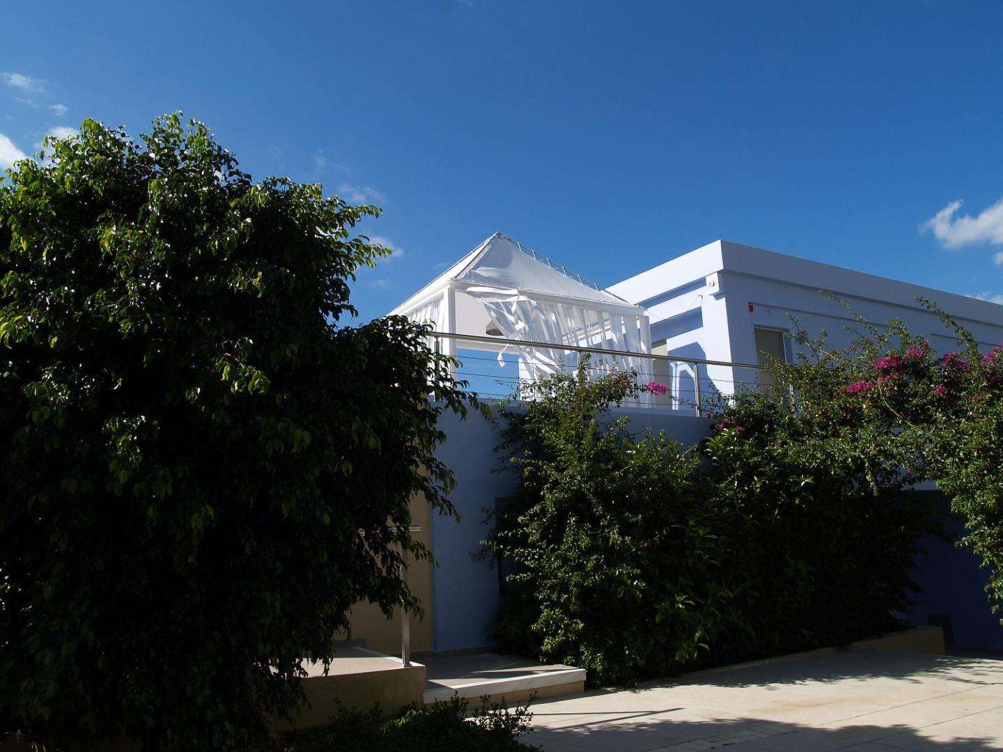 Mare Dei Ionian Resort modernās arhitektūras ēka ar terasi un baltu tentu, ko ieskauj zaļi krūmi un ziedoši augi