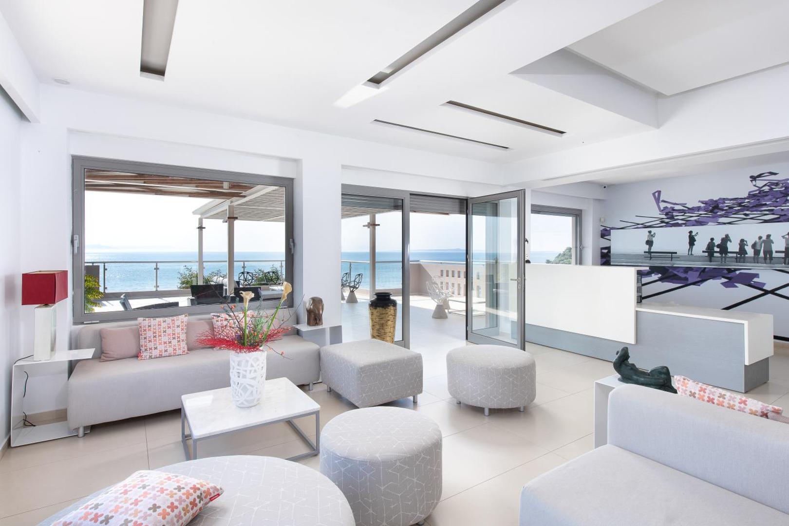 Mare Dei Ionian Resort gaišais lobby un atpūtas zona ar modernām mēbelēm, lieliem logiem un skatu uz Jonijas jūru.