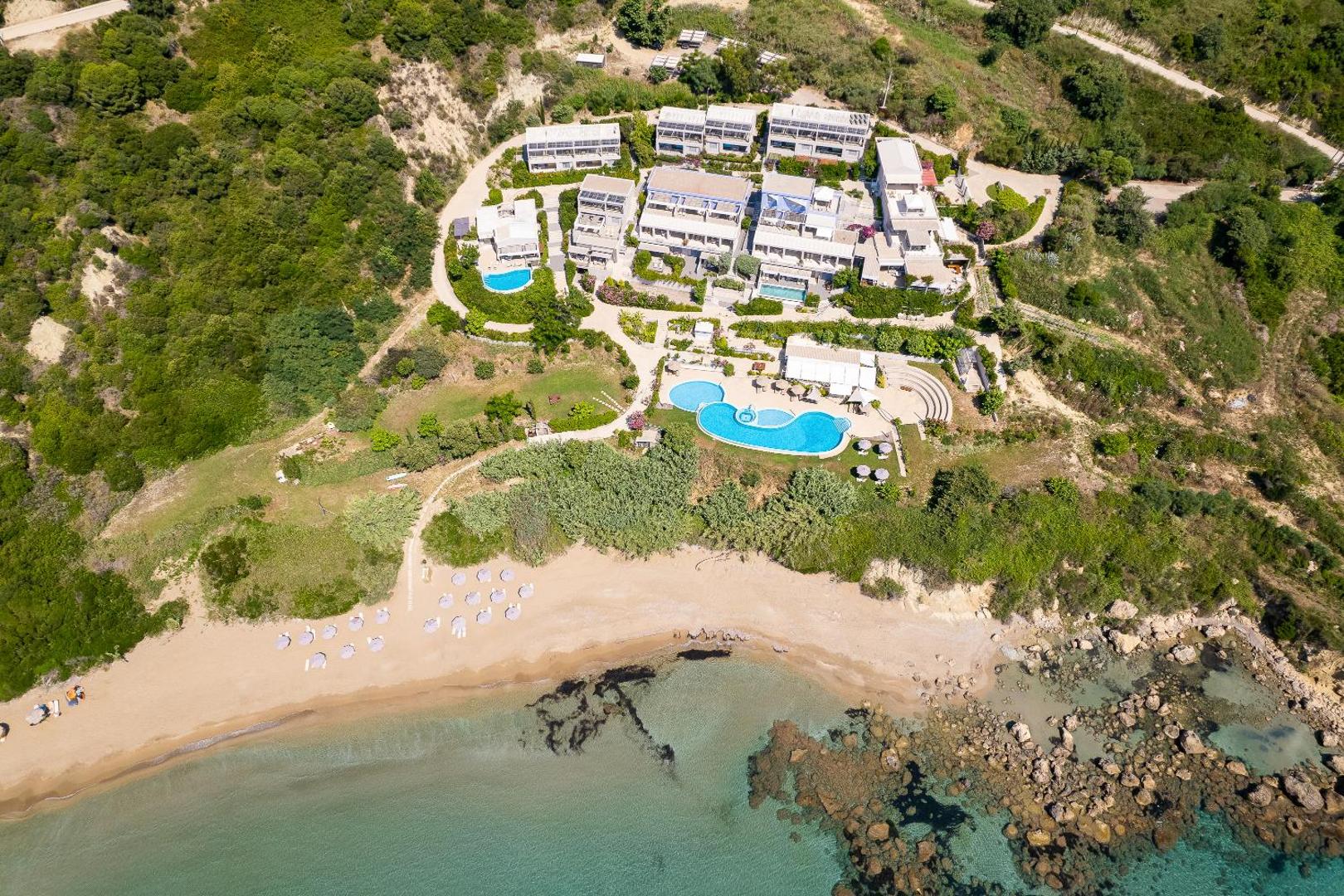 Mare Dei Ionian Resort viesnīcas teritorijas aerofoto, kurā redzami baseini, dārza celiņi un smilšains krasts ar akmeņiem ūdenī