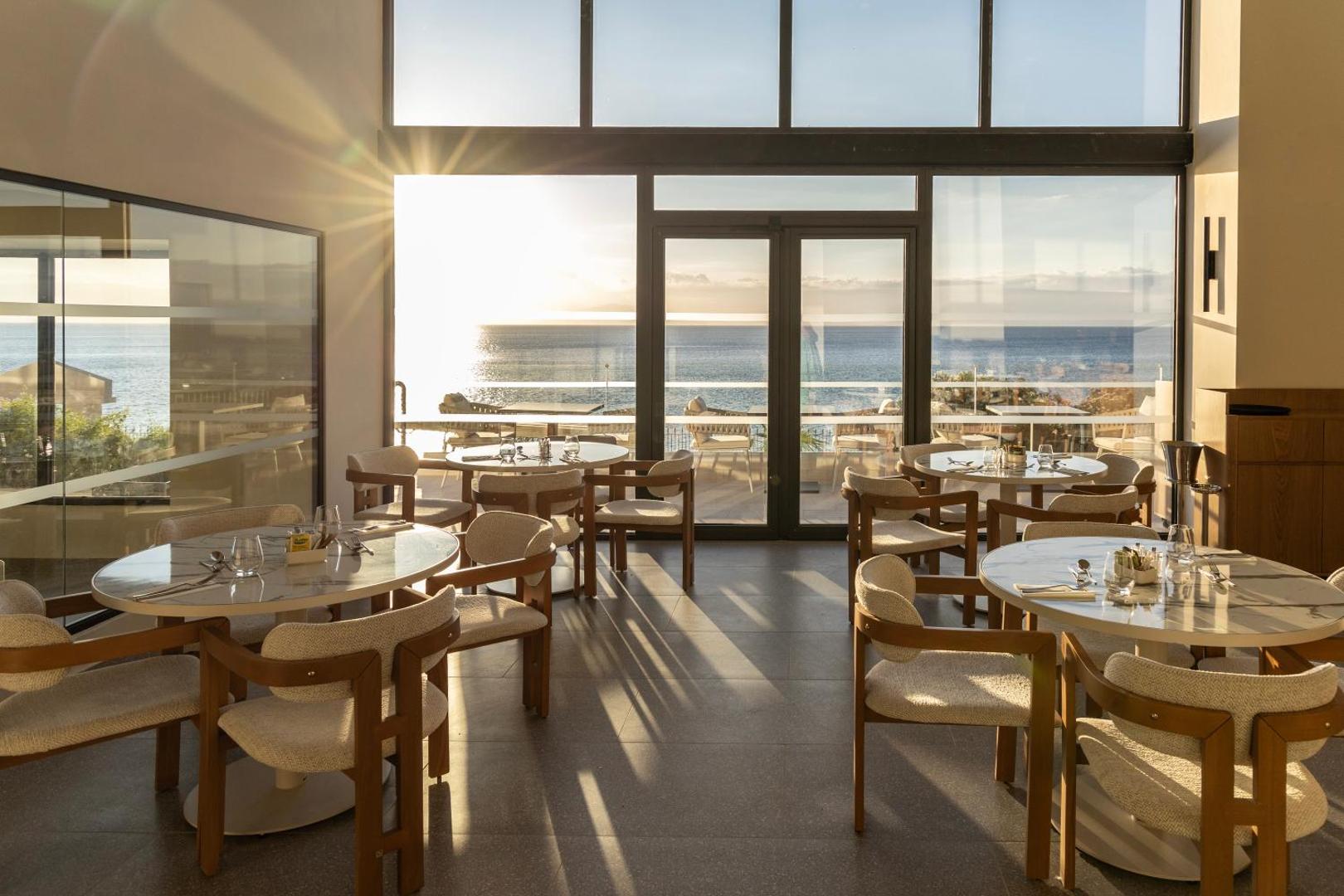 Valmar Corfu restorāna iekštelpas ar lieliem panorāmas logiem, kuros redzams saulriets pār jūru un eleganti klāti galdi