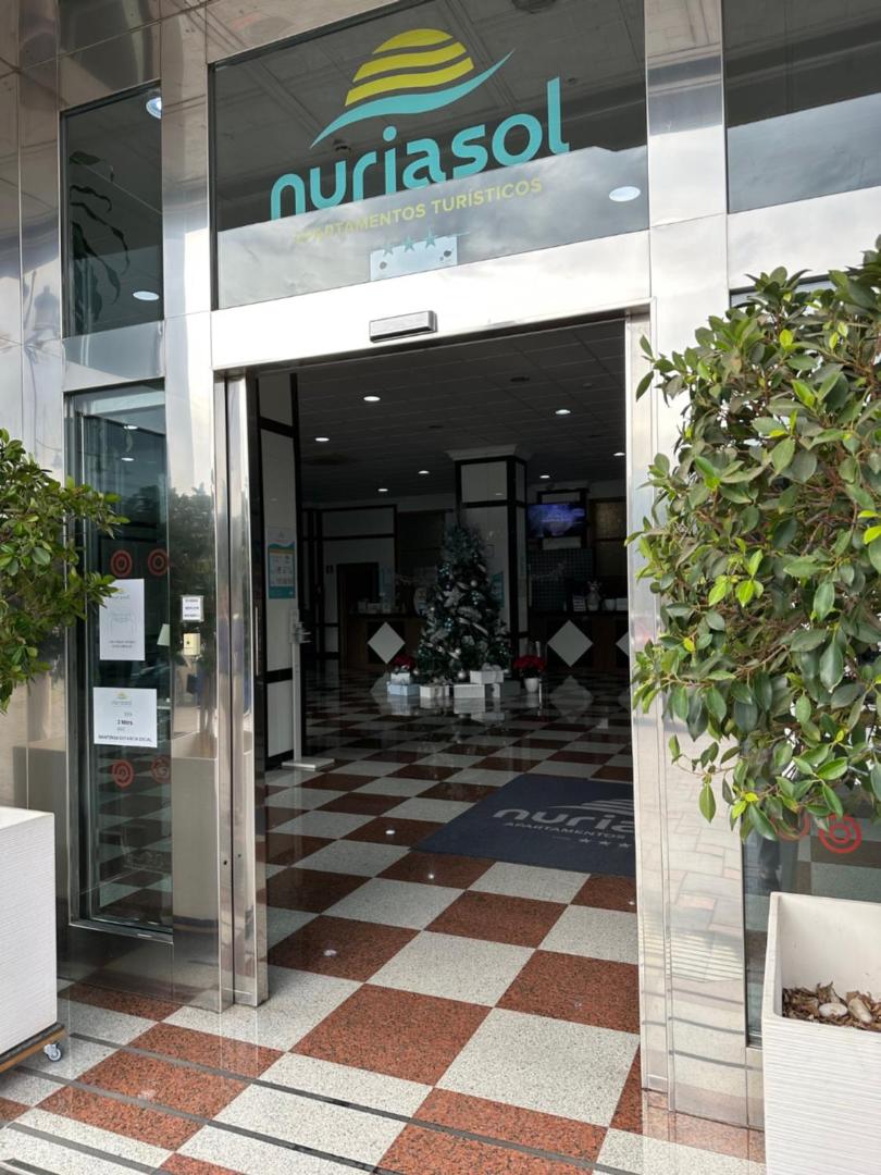 Apartamentos Nuria Sol ieeja ar stikla durvīm un viesnīcas logo Fuengirolā, Spānijā — skats uz vestibilu ar dambretes stila grīdu