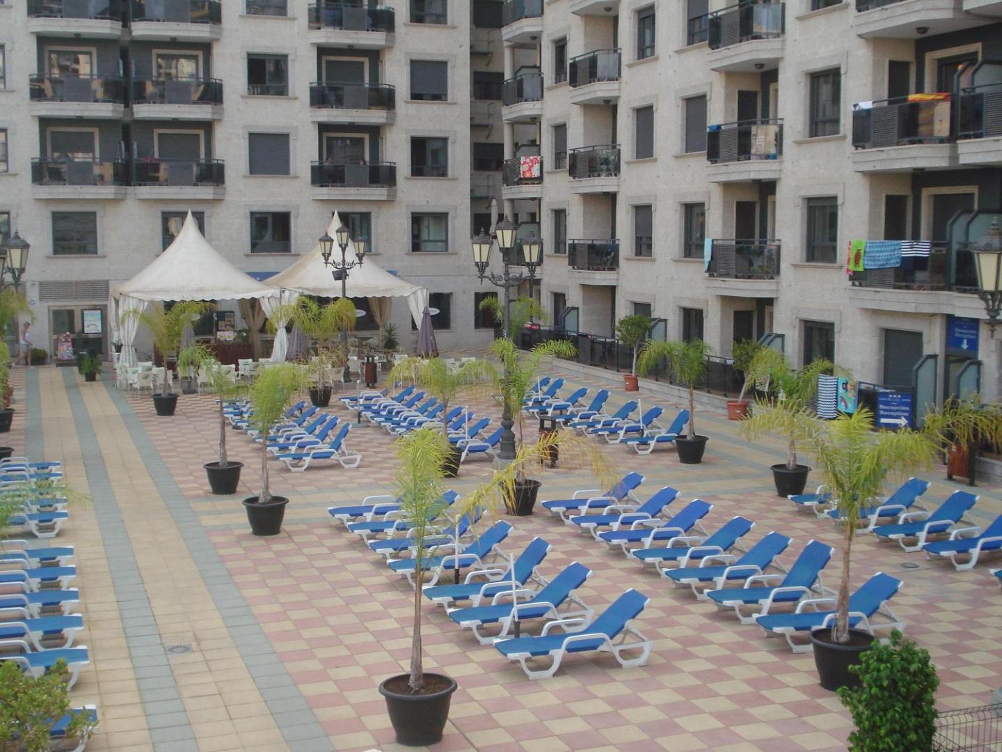 Apartamentos Nuria Sol iekšpagalms ar daudziem ziliem sauļošanās krēsliem un dekoratīvām palmām podos starp viesnīcas korpusiem