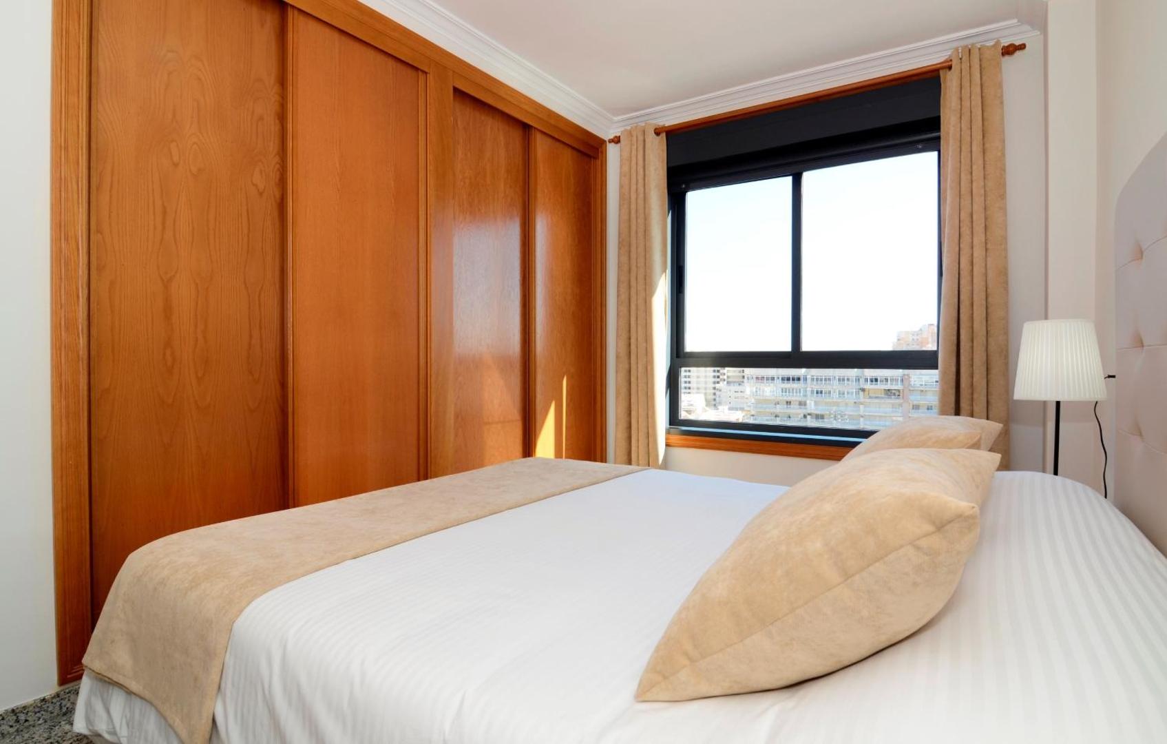 Apartamentos Nuria Sol guļamistabas interjers ar gaišu gultasveļu, koka skapi un logu, pa kuru ieplūst dabiskā gaisma