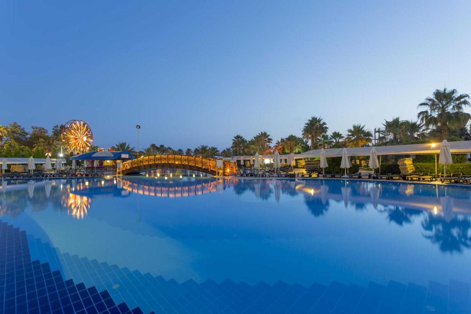 Mukarnas Spa & Resort plašā baseina zona krēslas stundā ar skatu uz panorāmas ratu un palmu dārzu