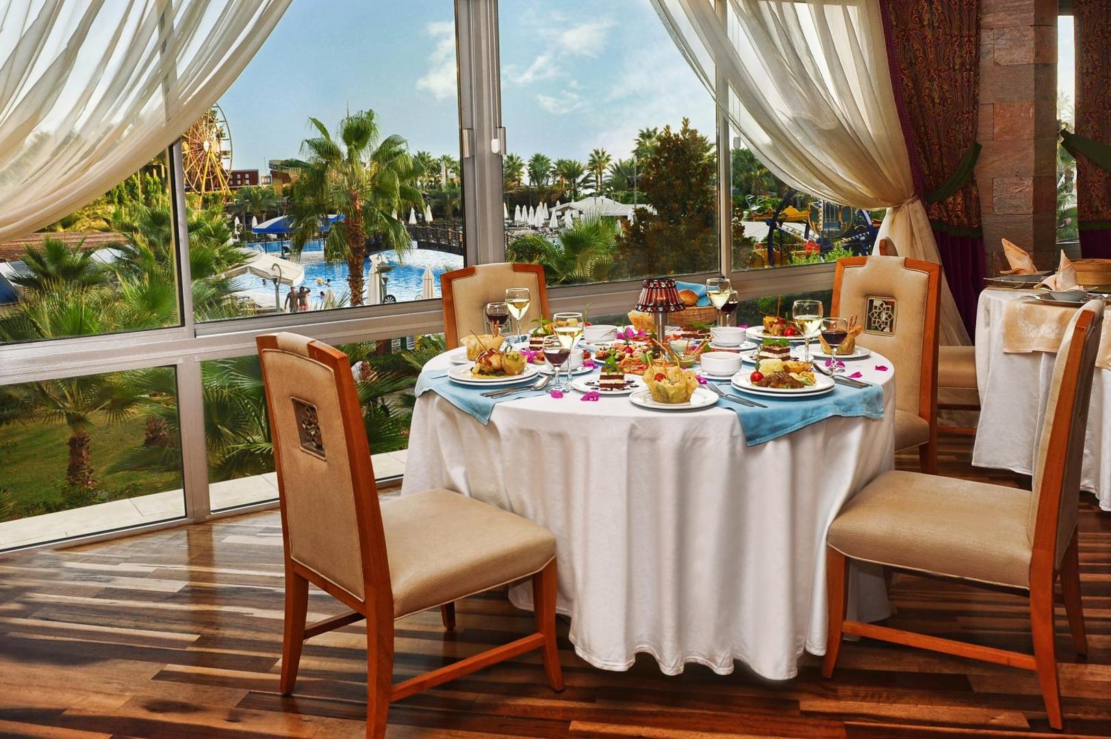 Mukarnas Spa & Resort restorāna galda klājums ar skatu pa lielu logu uz viesnīcas dārzu, baseiniem un panorāmas ratu