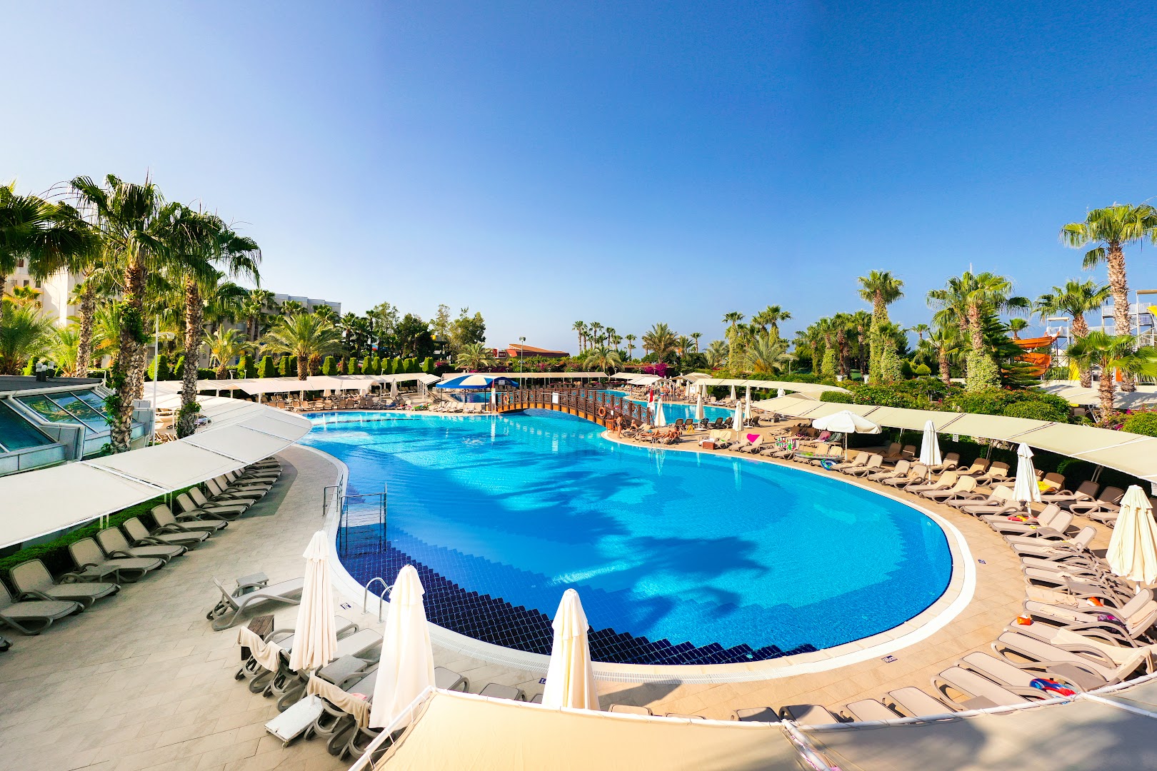 Mukarnas Spa & Resort lielais āra baseins saulainā dienā ar daudziem sauļošanās krēsliem, saulessargiem un palmām