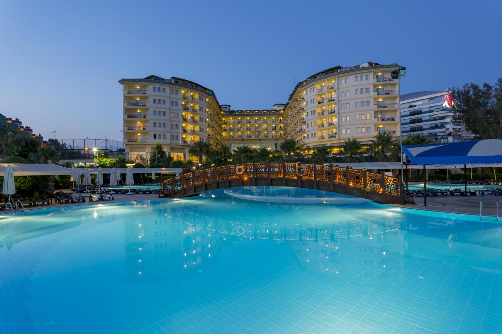 Mukarnas Spa & Resort viesnīcas ēka un izgaismots āra baseins ar koka tiltiņu vakarā Alanjas reģionā, Turcijā