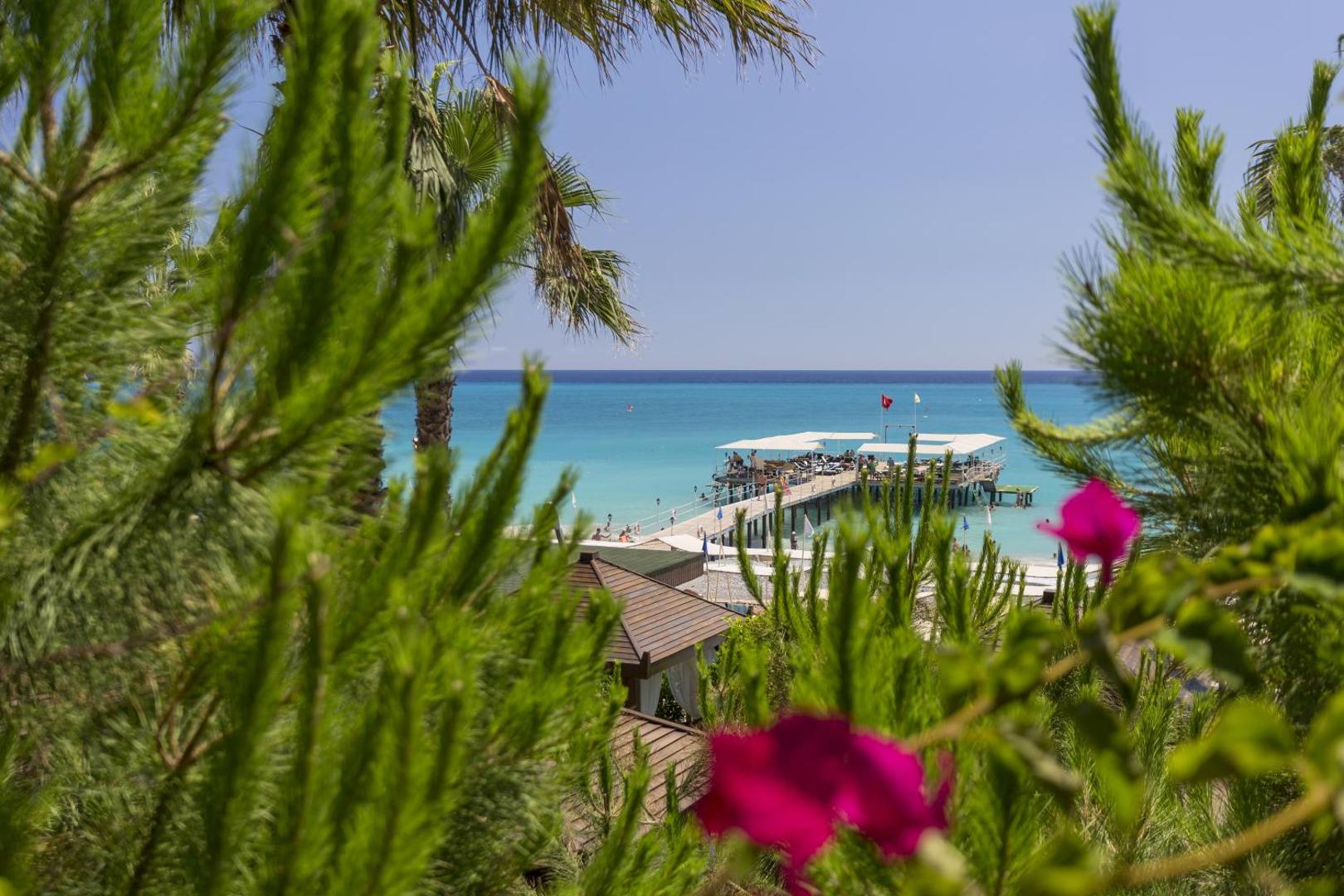 Mukarnas Spa & Resort skats uz Vidusjūru caur zaļojošiem kokiem — tālumā redzama koka piestātne ar atpūtas zonu un tirkīzzils ūdens