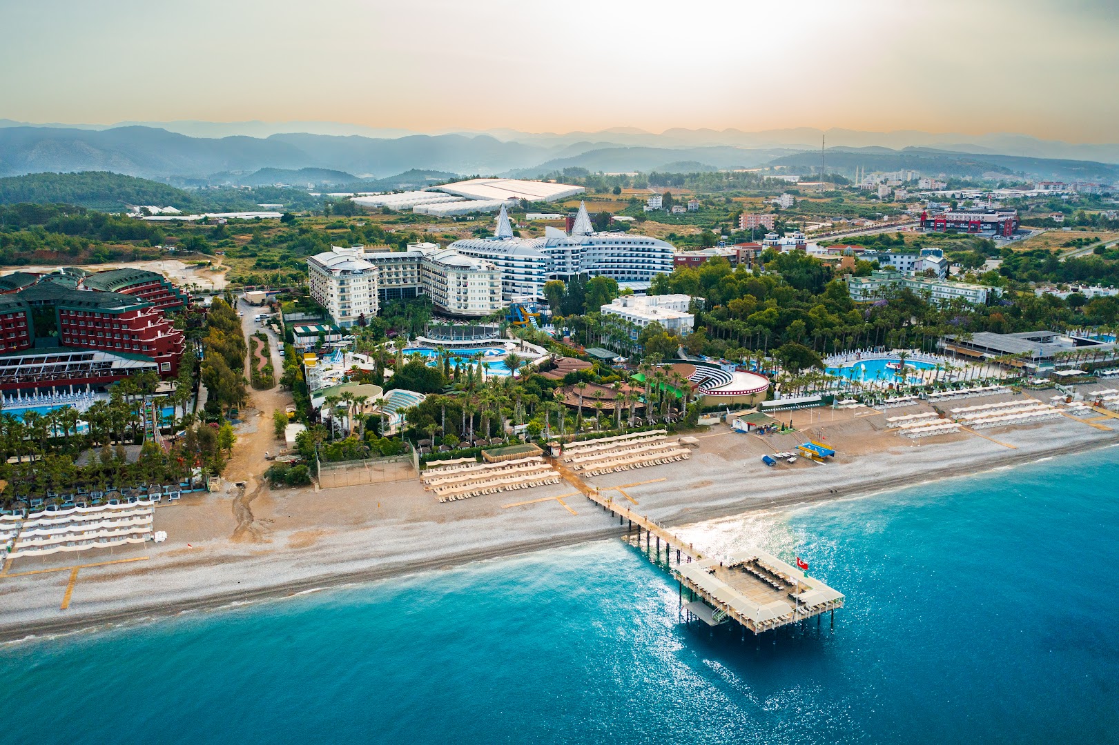 Mukarnas Spa & Resort panorāmas skats no putna lidojuma — viesnīcas ēkas, plaša pludmale, piestātne un baseini Alanjas piekrastē