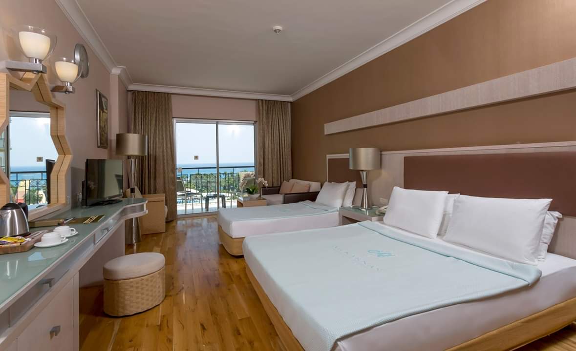 Mukarnas Spa & Resort numura interjers ar divām gultām, tualetes galdiņu un plašu stikloto sienu uz balkonu ar skatu uz jūru